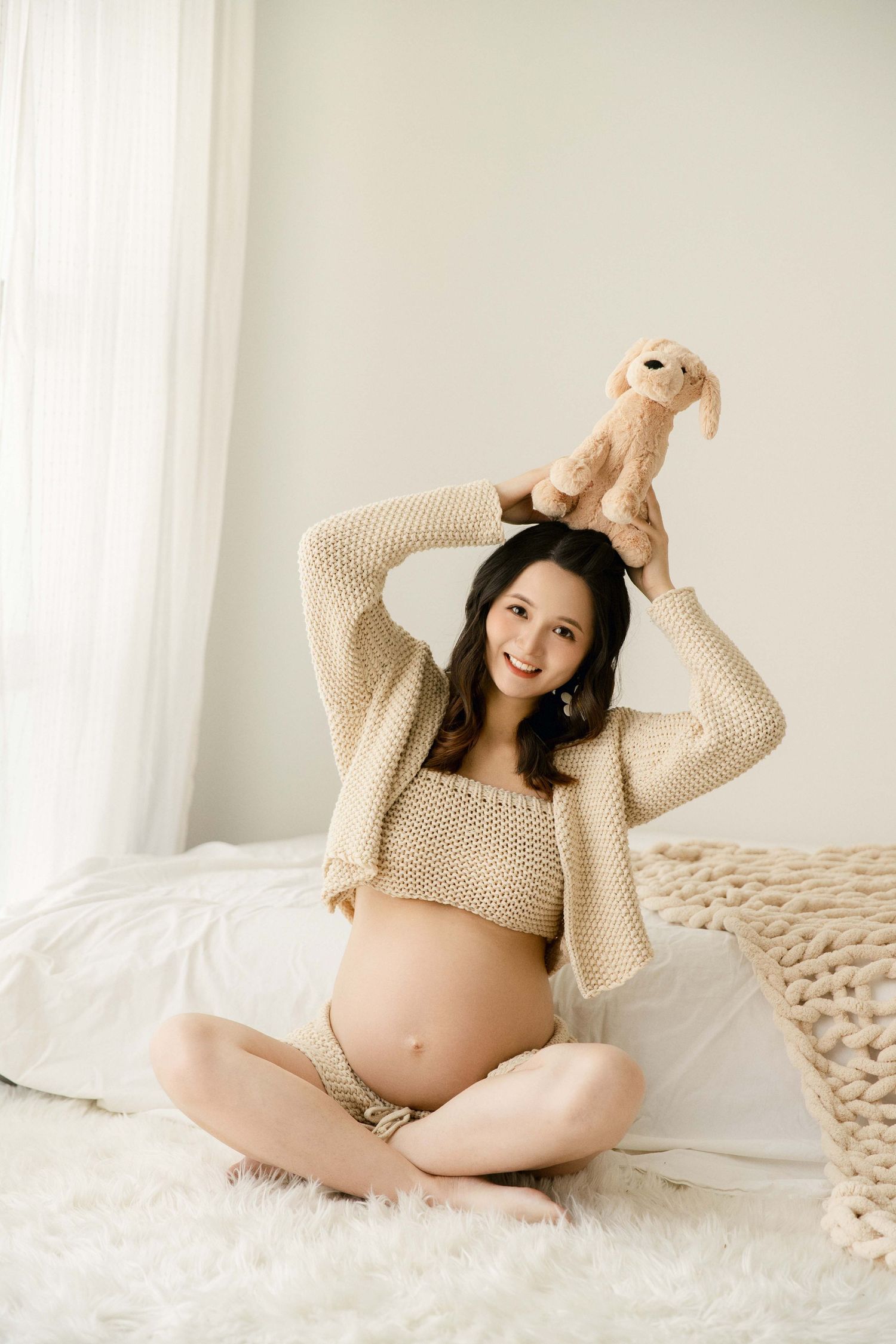 Playful maternity photos with stuffed animal and cream knit sweater on textured bedding.