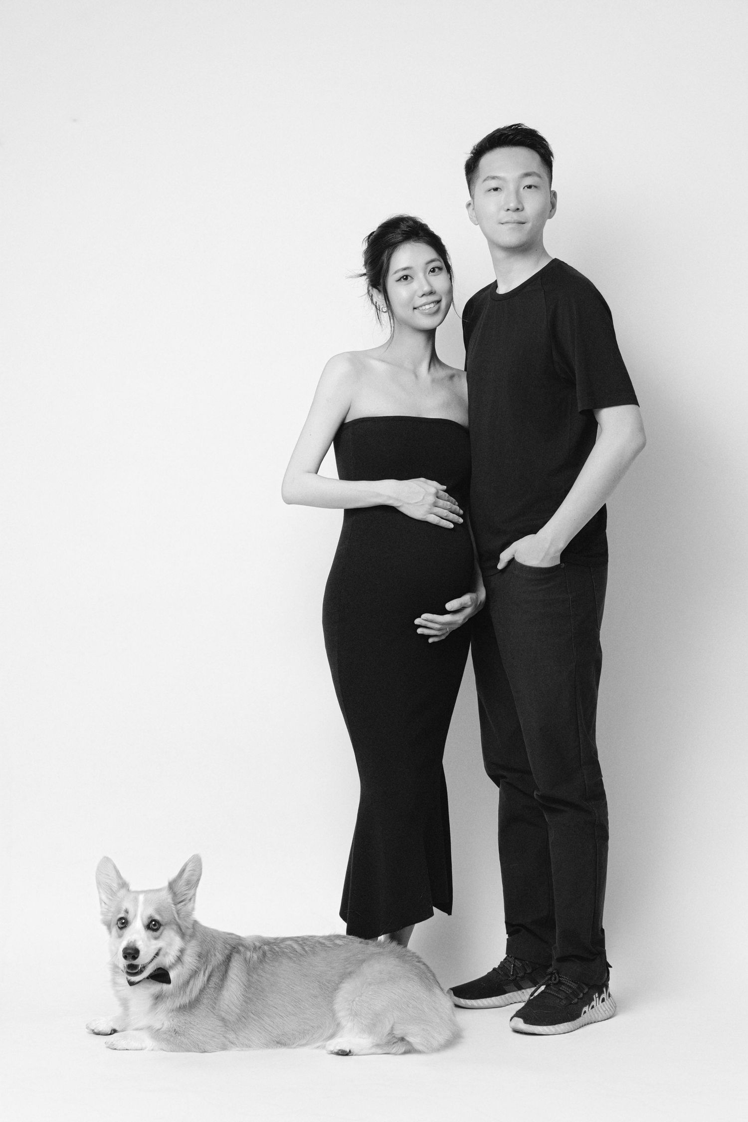 Black and white maternity photo with couple and Corgi dog in minimalist studio setting.