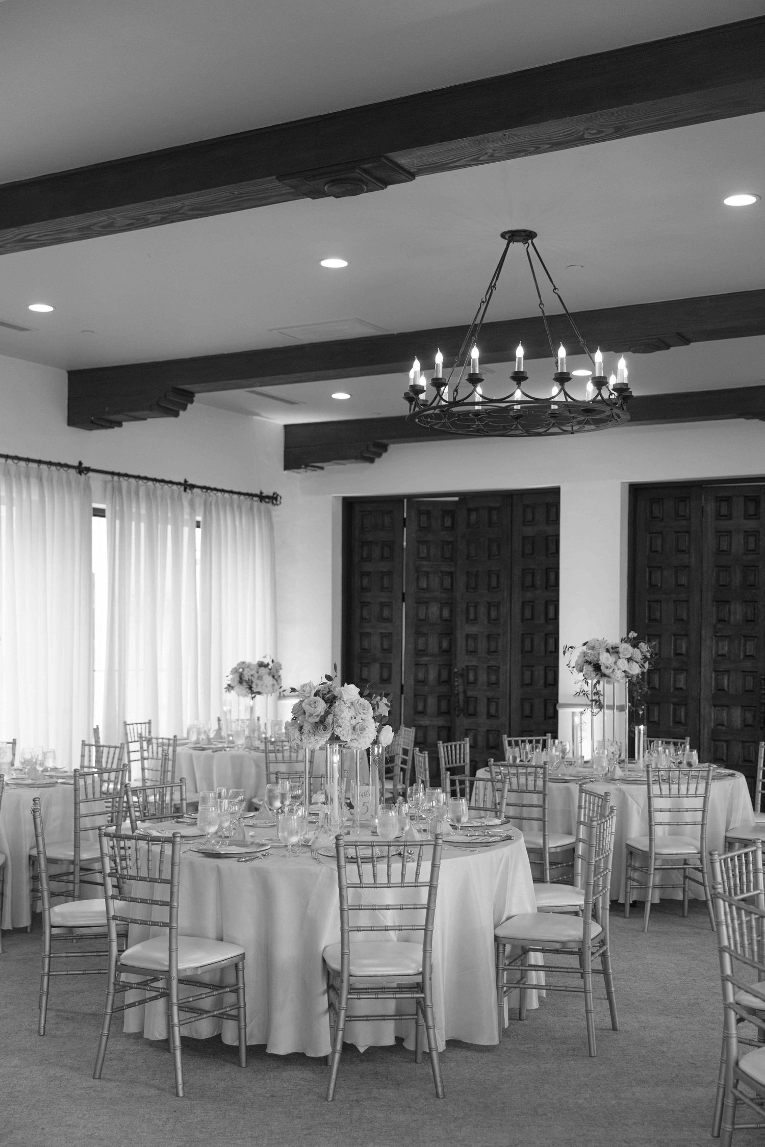 Elegant wedding reception venue with round tables, silver chiavari chairs, and a black beam ceiling with chandelier.