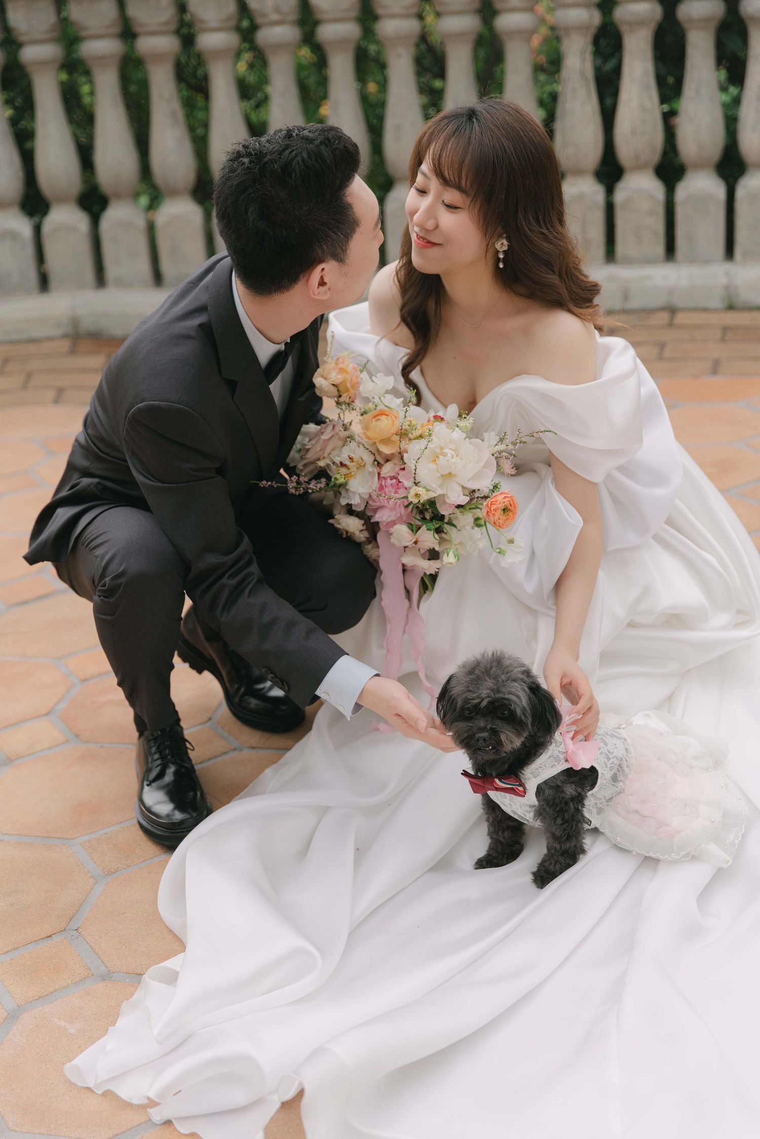 Wedding couple poses with their small black dog on a terrace with floral bouquet.