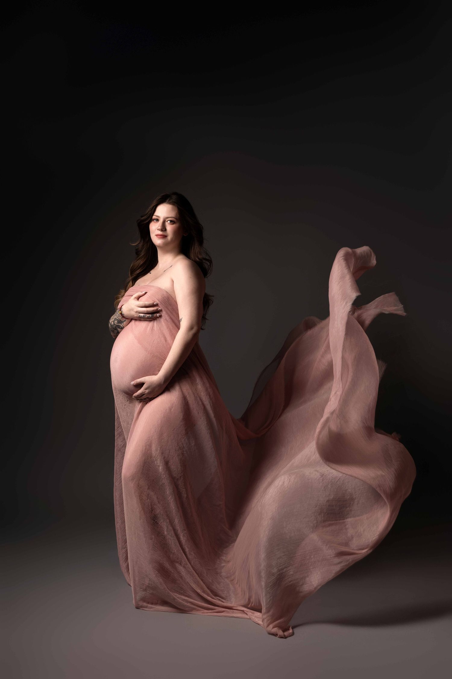 Dramatic maternity photography with flowing pink dress against dark background.