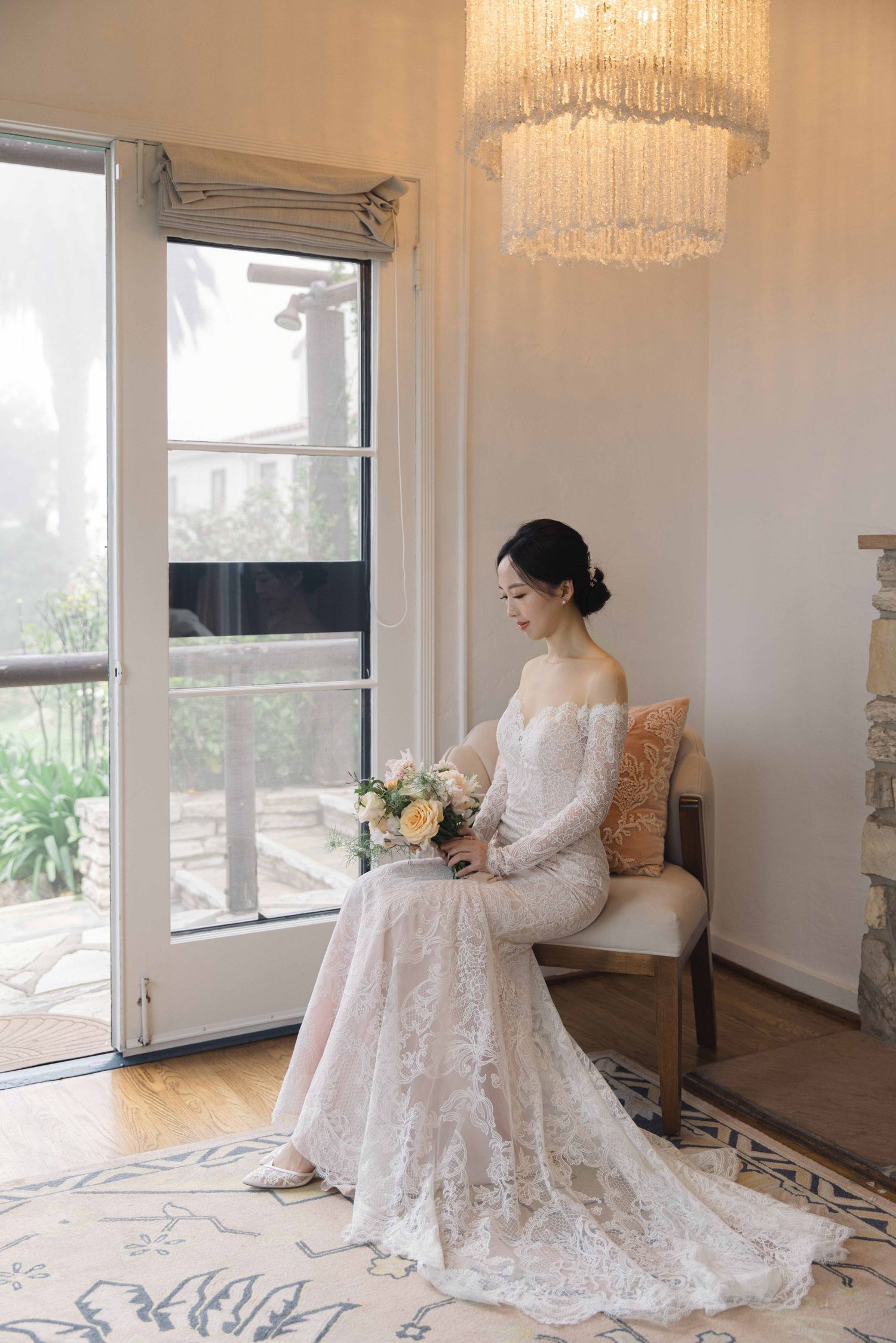 A bride in an elegant white lace gown sits gracefully on a chair near large windows with crystal chandeliers.