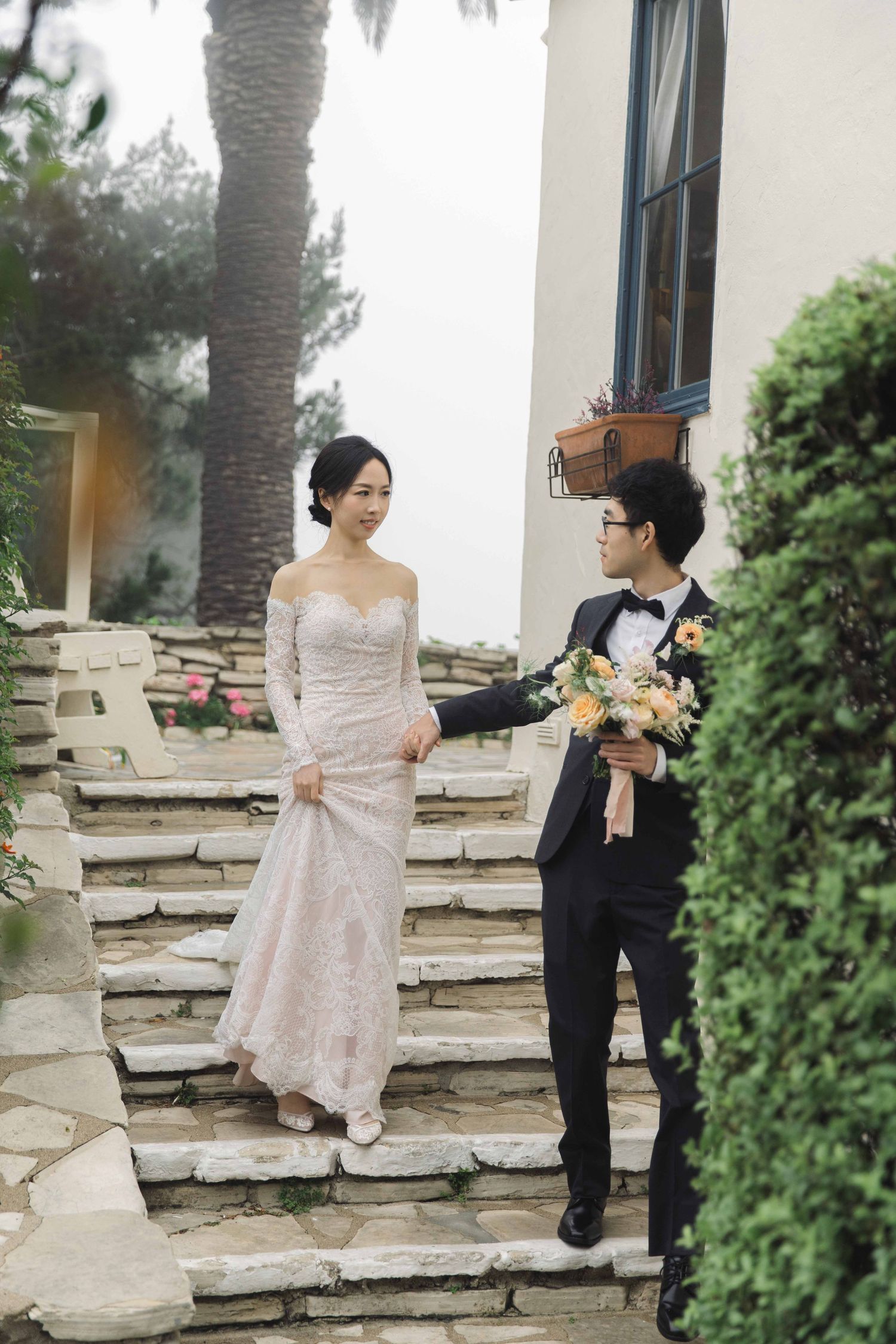 A wedding couple poses on stone steps surrounded by lush garden landscaping.
