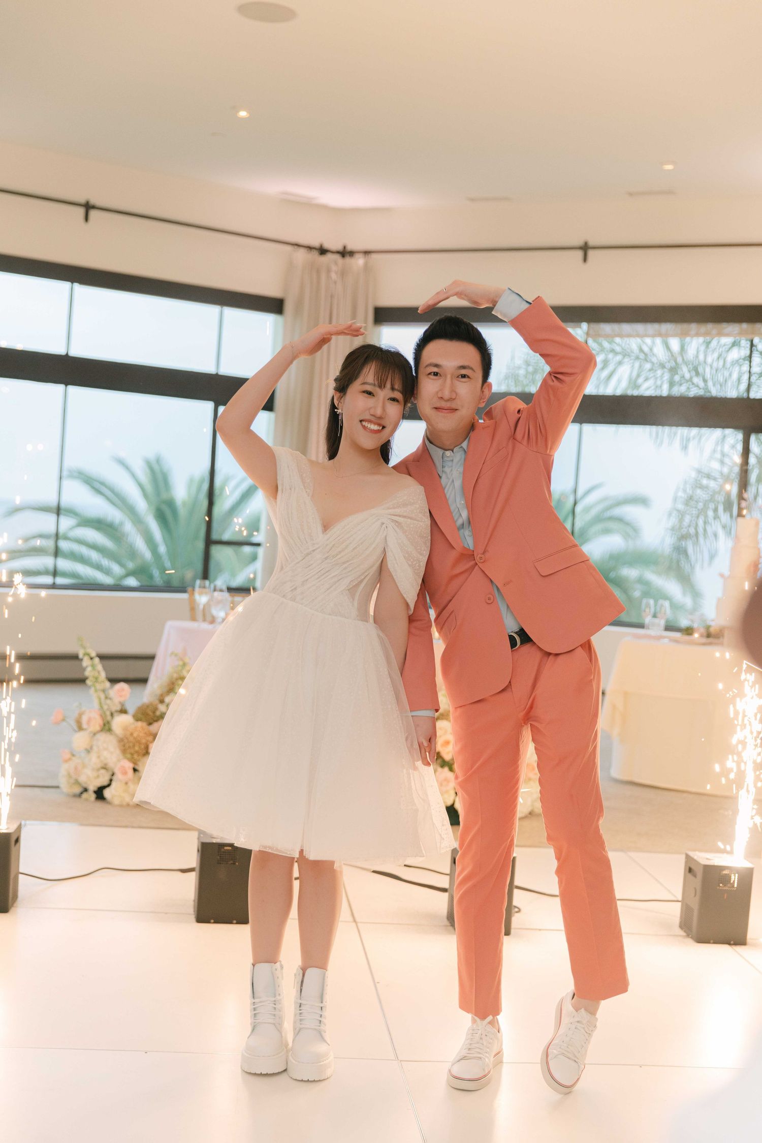 A couple in wedding attire dances playfully in a bright indoor space with palm trees visible outside.