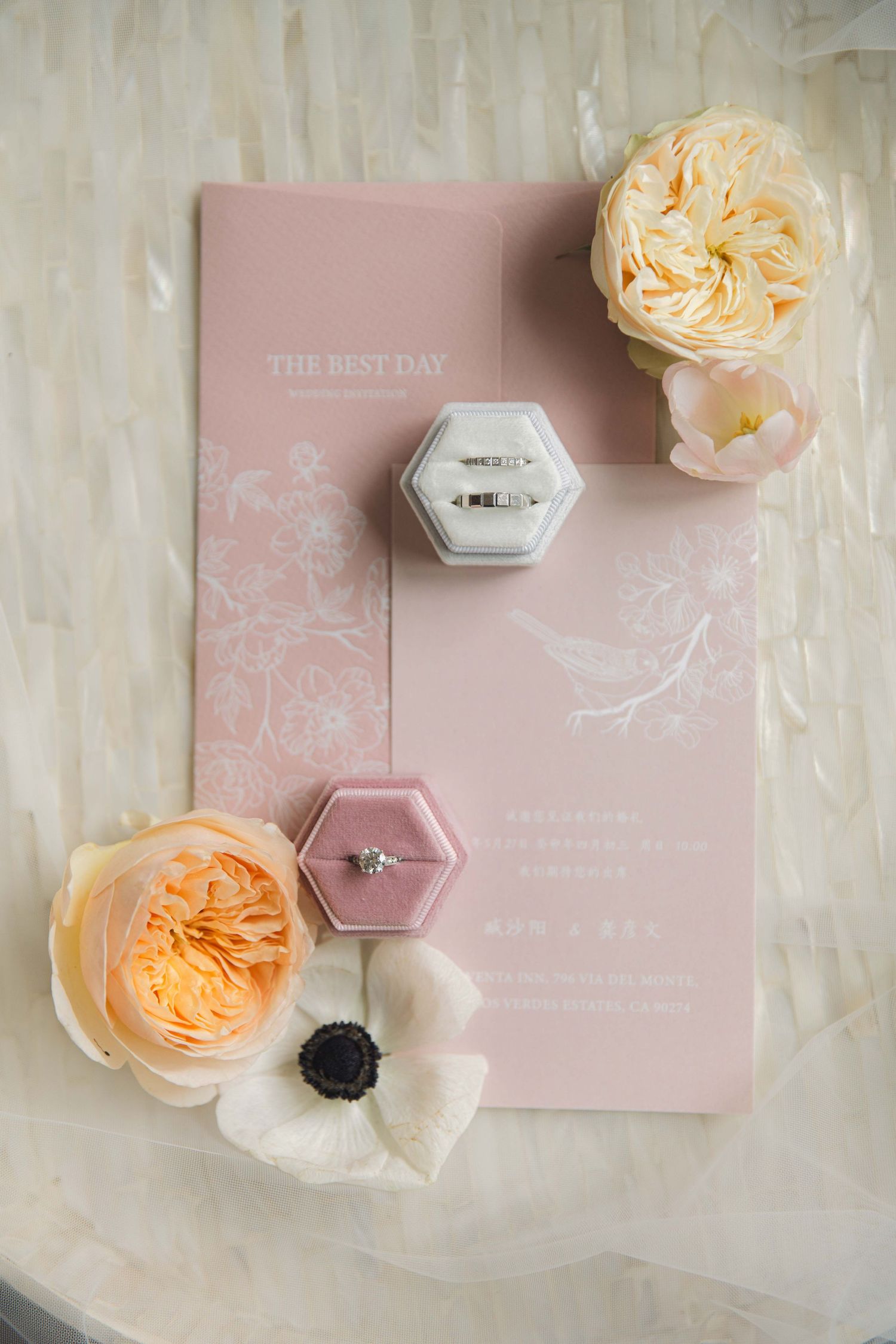 Pink wedding invitation with hexagonal ring boxes and peach garden roses on white fabric.