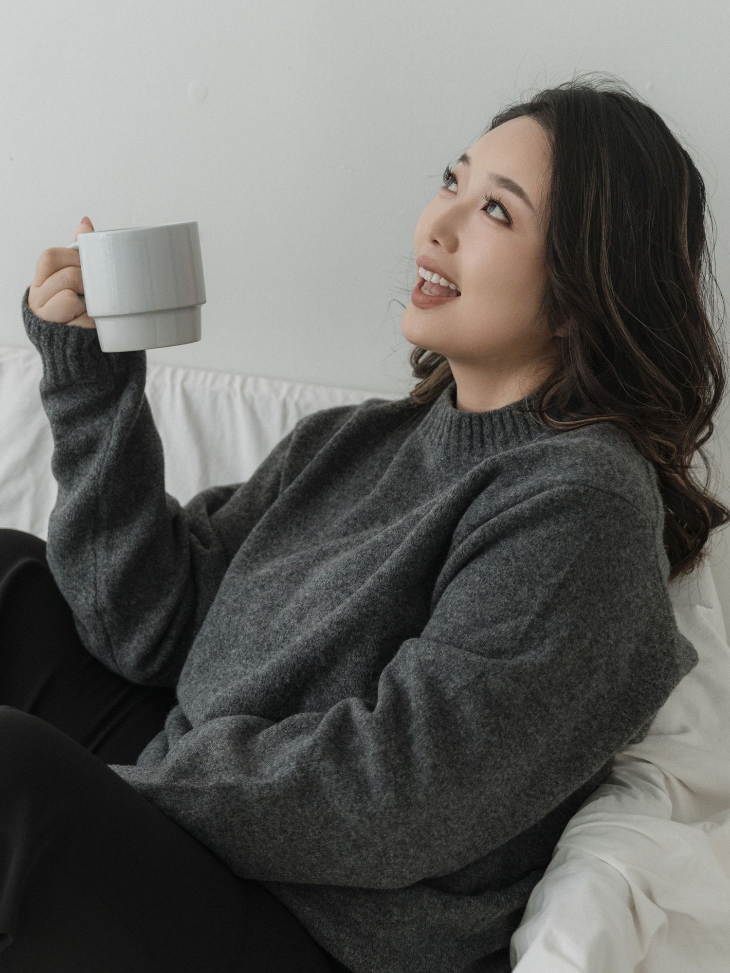 Person relaxing on white bedding while drinking from a mug in gray sweater.