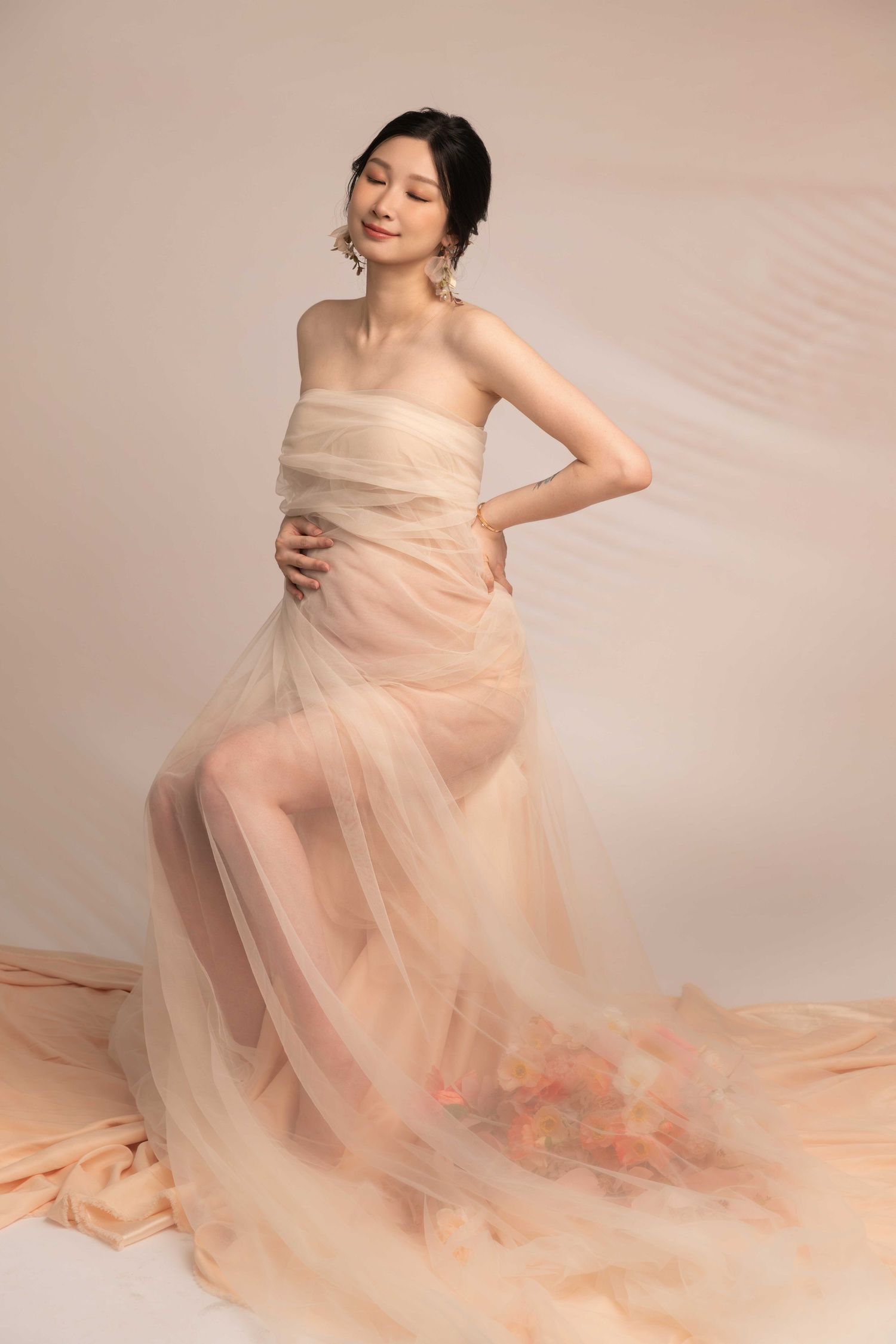 Romantic maternity session with flowing peach tulle gown in soft studio lighting.
