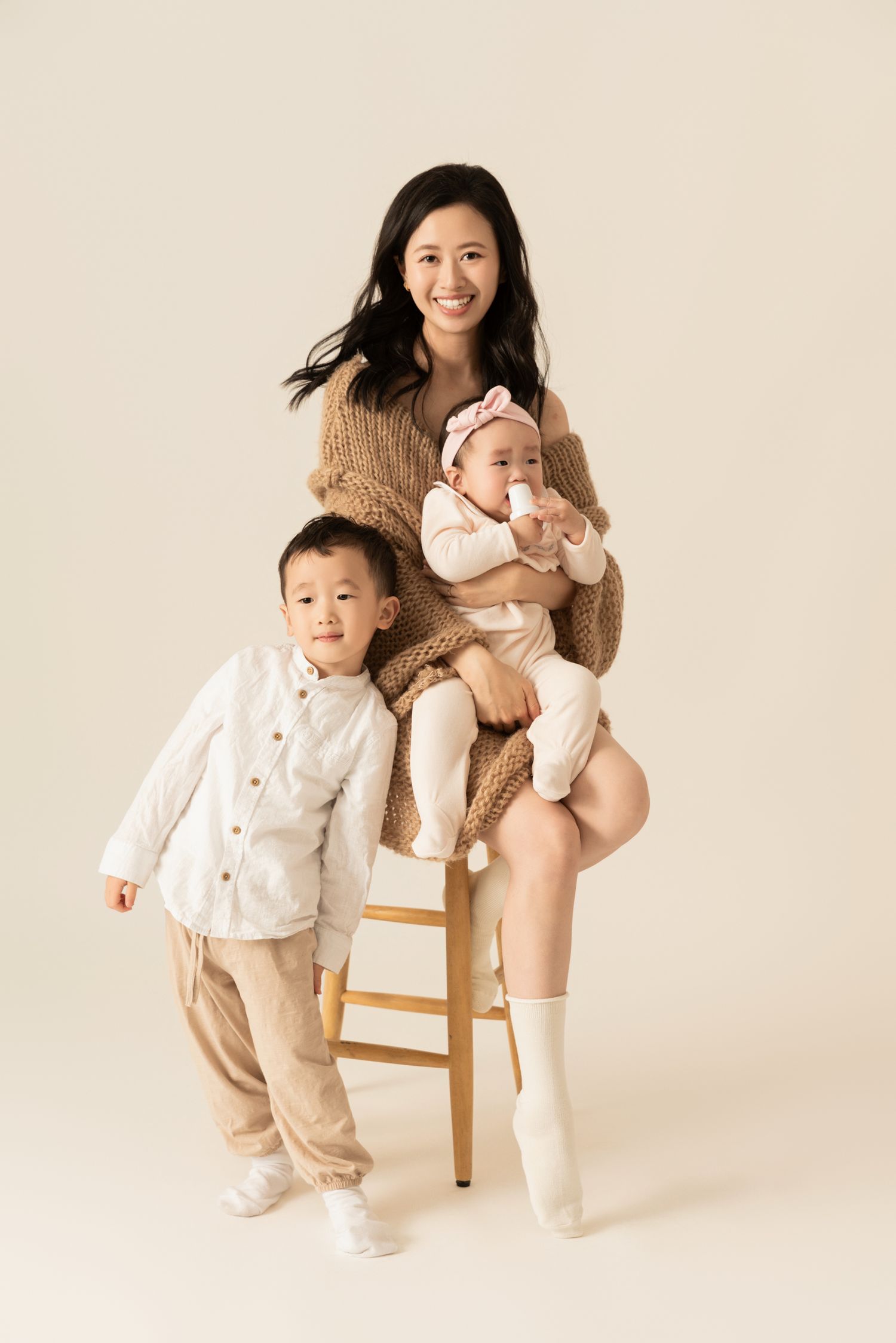 Portrait on wooden stool against neutral backdrop wearing coordinated cream and tan outfits.