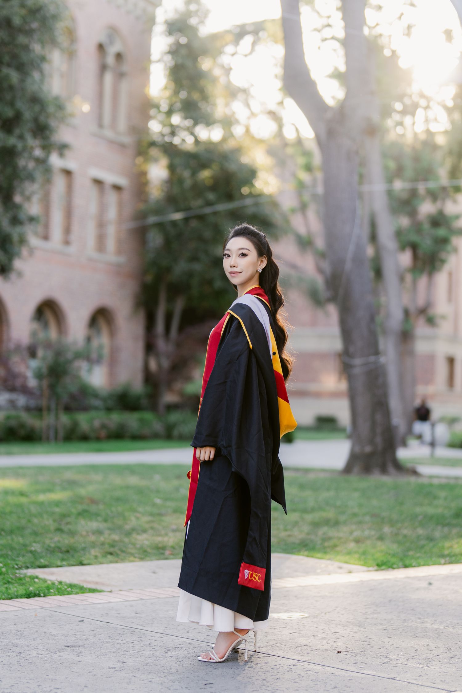 Graduate in black academic regalia walks along campus pathway during sunset.