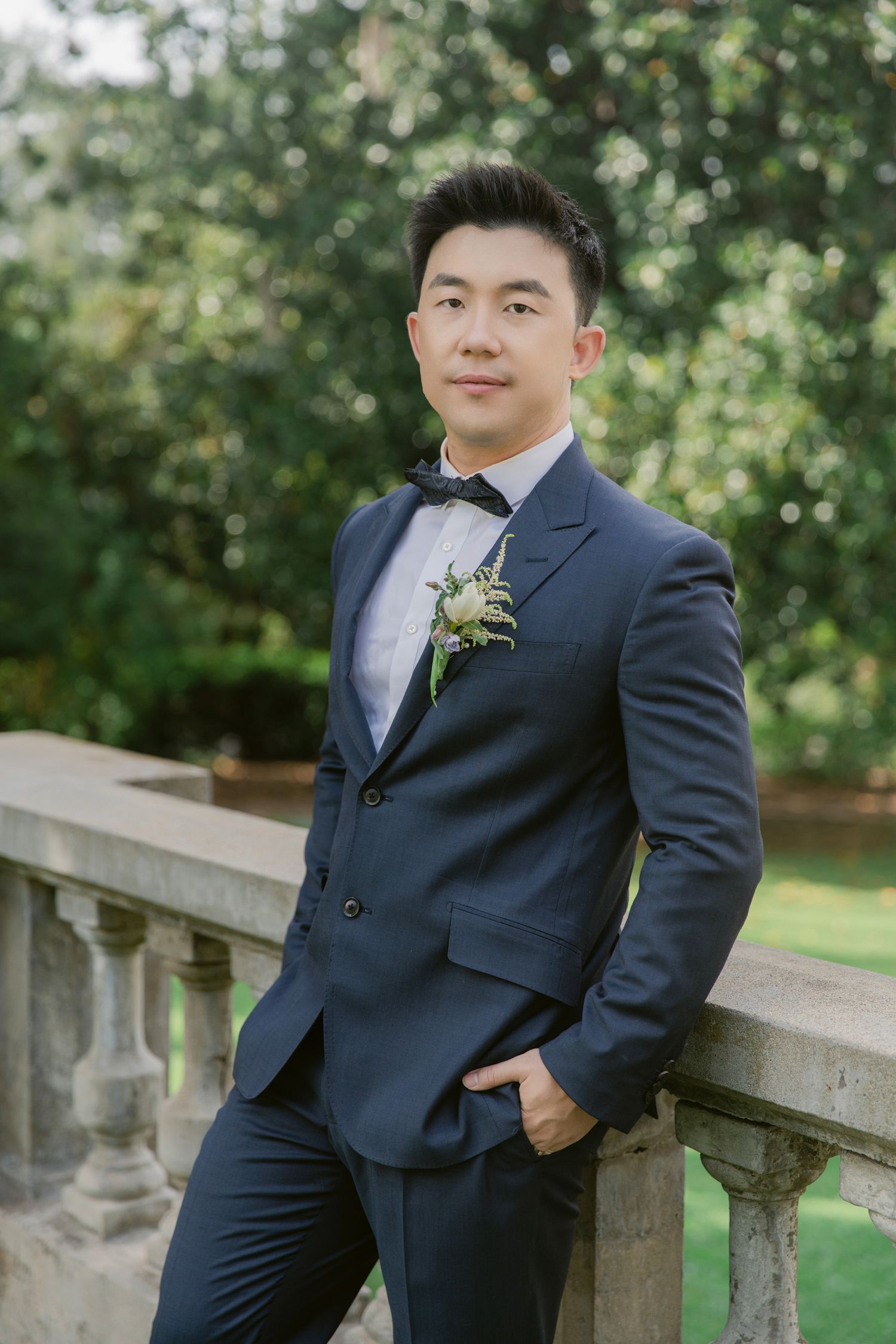 Groom in classic navy suit poses against stone balustrade in garden setting.