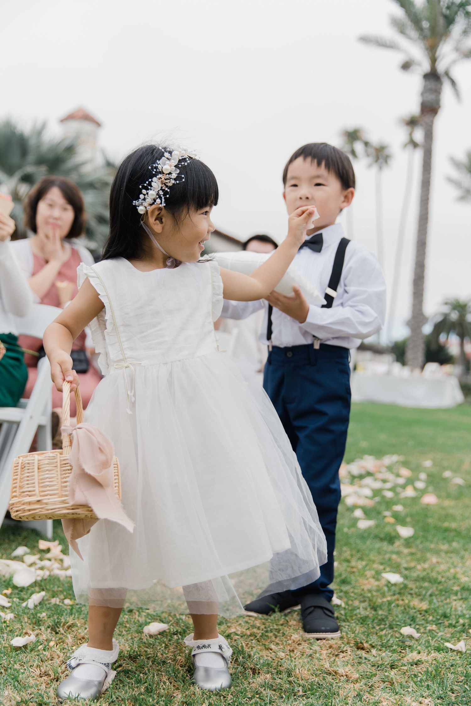 Young flower girl in white dress plays during wedding ceremony on grass lawn.
