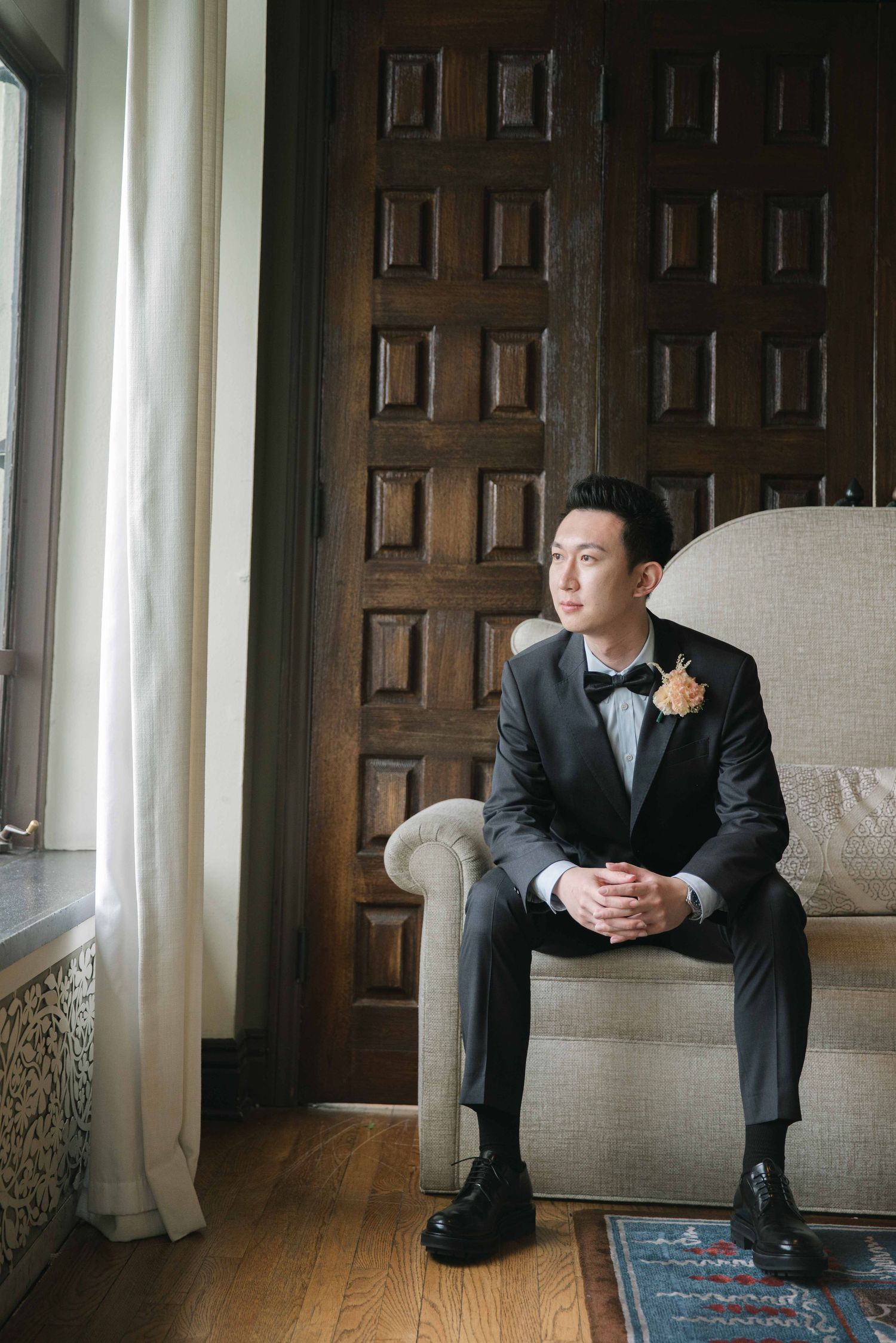 A formal wedding portrait session featuring a groom in a black suit sitting thoughtfully by a window.