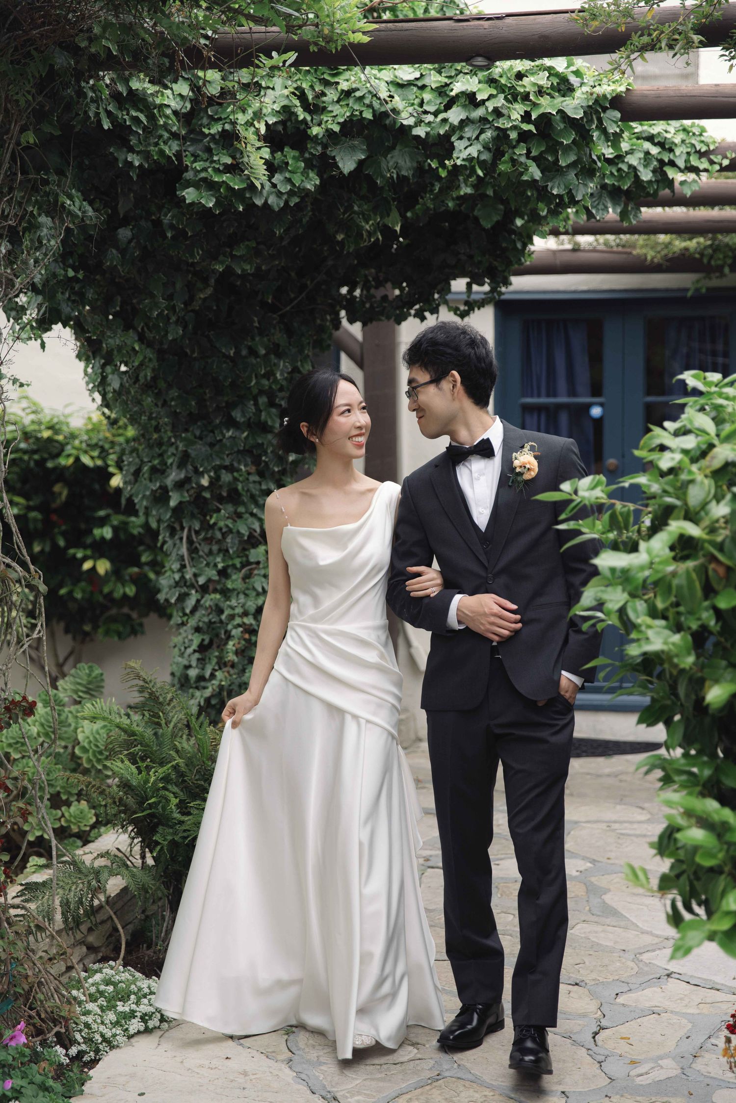 A couple walks along a vine-covered garden path in elegant wedding attire.