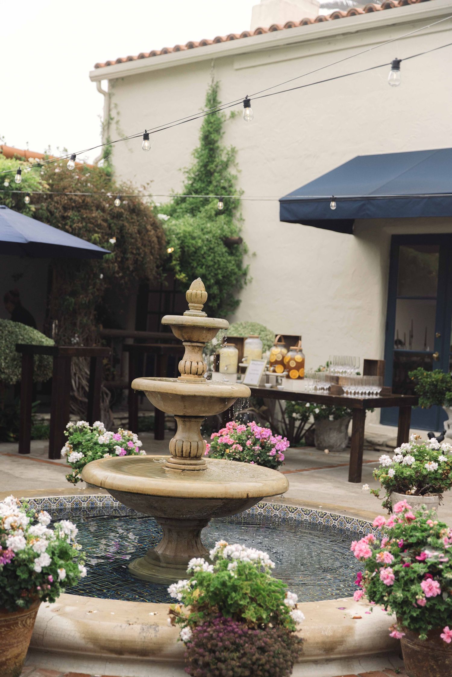 Elegant garden fountain surrounded by pink flowers and potted plants on a stone patio.