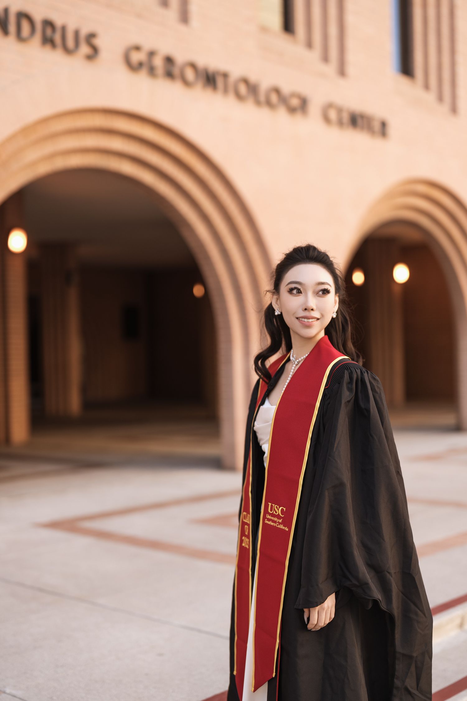 Graduate in black gown and red stole posing at campus building entrance.