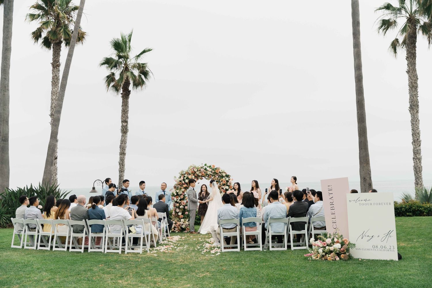 Wedding ceremony setup with white chairs and floral arch under tall palm trees.