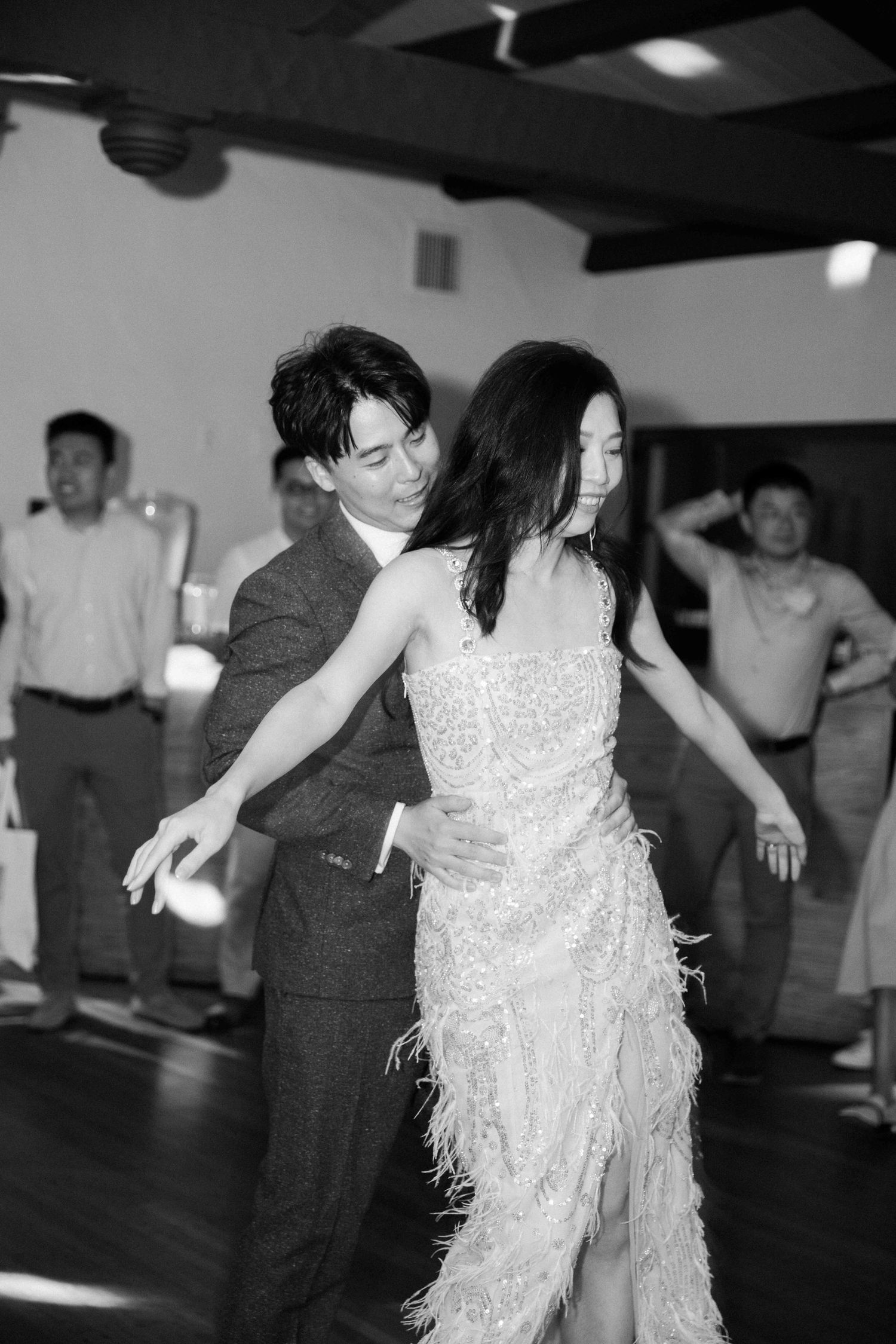 People dancing in formal attire at a wedding reception with a white dress visible in the frame.