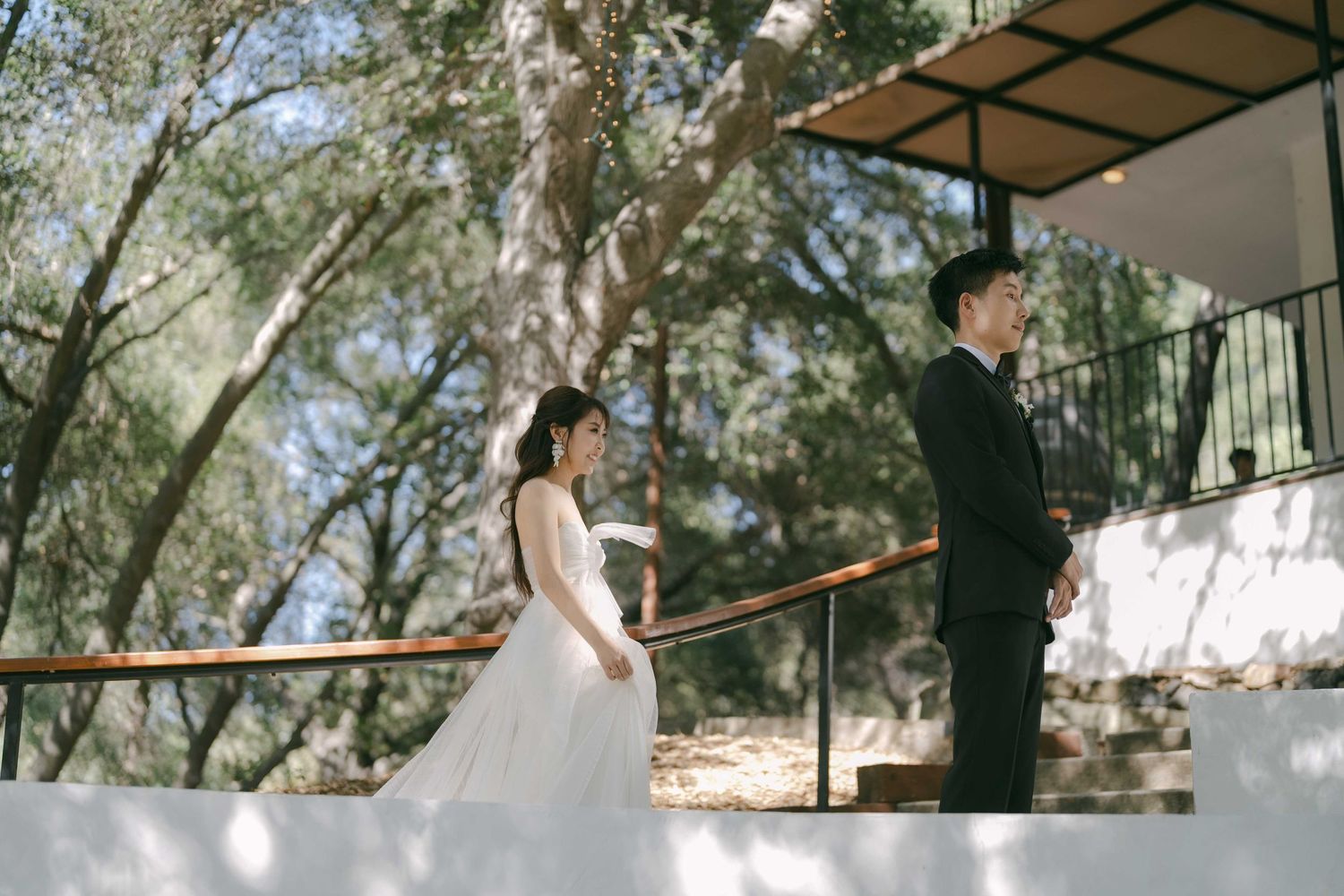 Outdoor wedding ceremony setup with trees and white decorative elements in natural lighting.