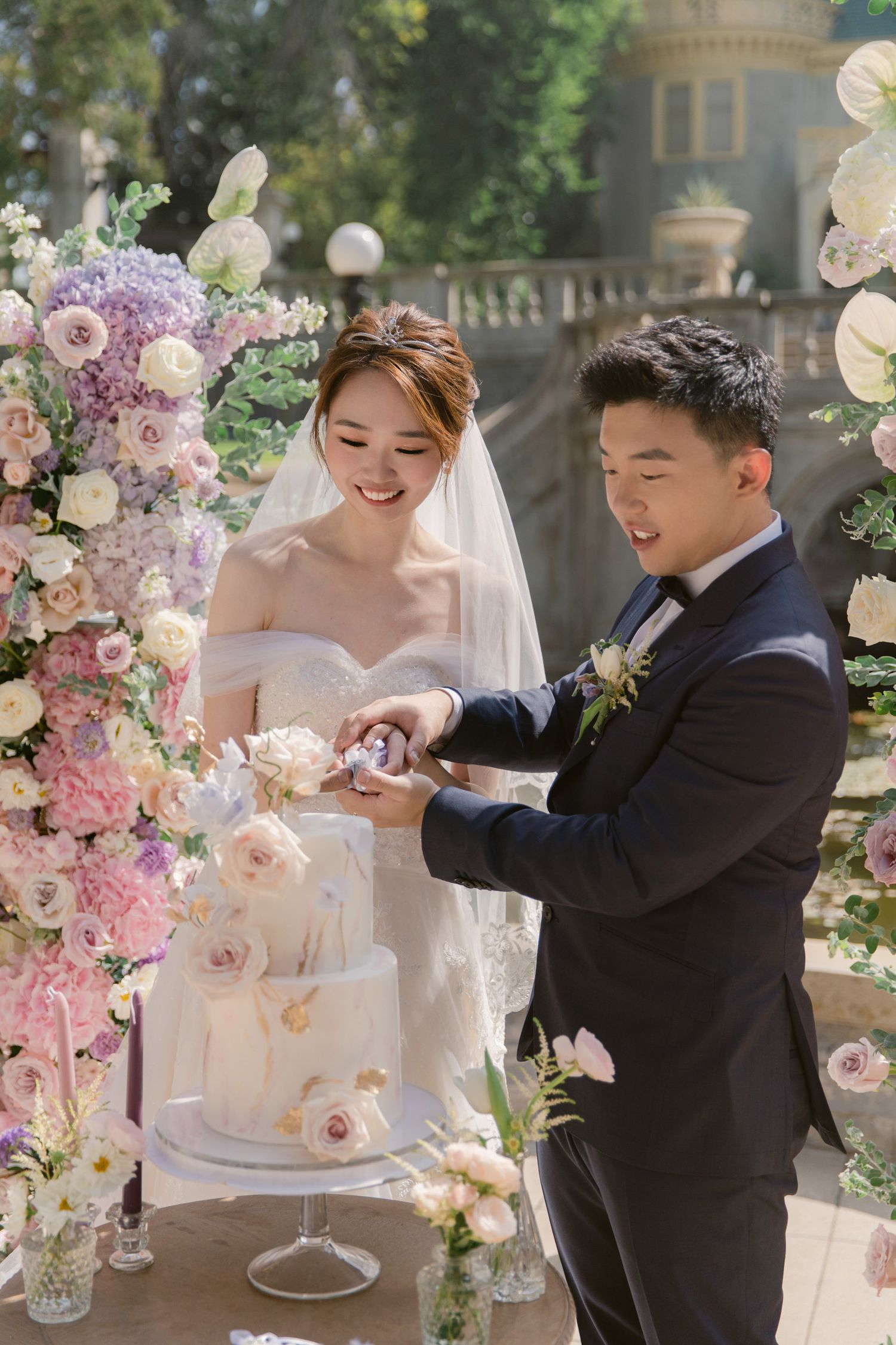 Elegant wedding cake cutting ceremony with delicate pink floral decorations in an outdoor garden setting.