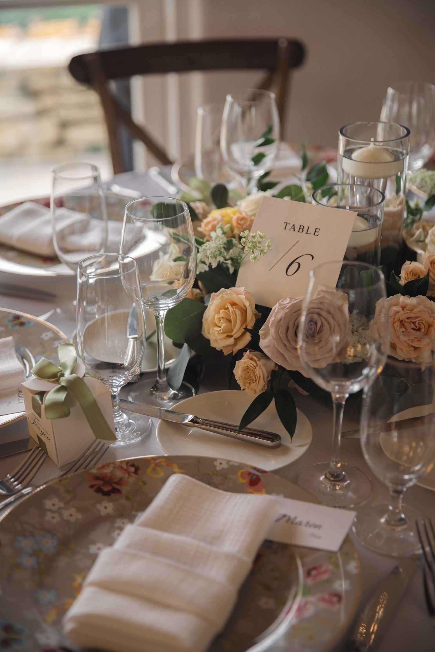 Wedding table setting with peach roses, table number 6, and crystal glassware on floral patterned plates.