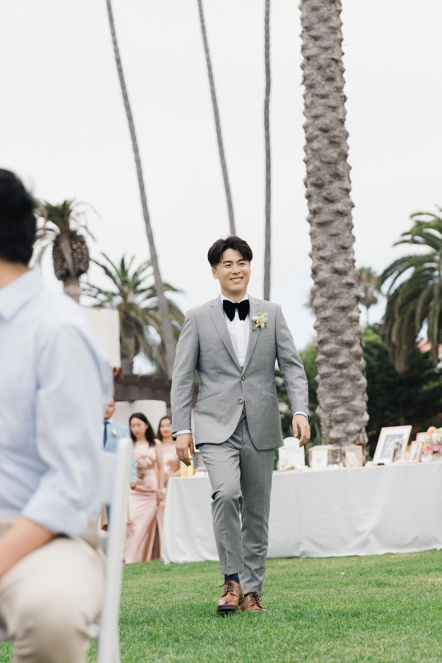 Wedding ceremony guest walks down a palm tree-lined outdoor aisle in a grey suit.