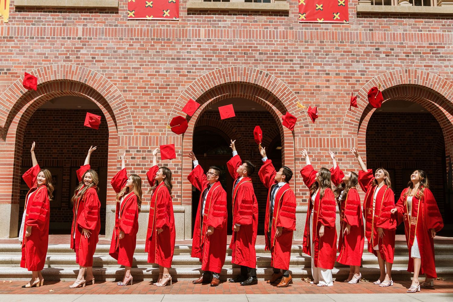 Graduates in red caps and gowns pose in sequence along brick university building facade.