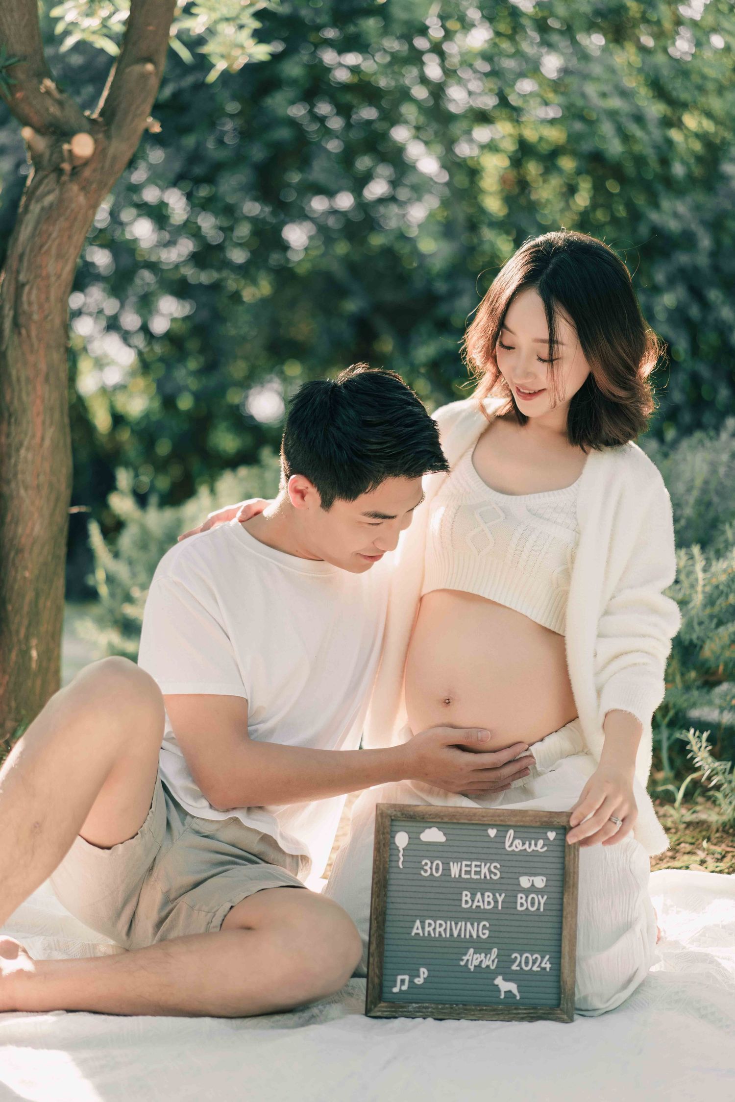 Outdoor maternity photoshoot with chalkboard baby announcement and couple on grass.