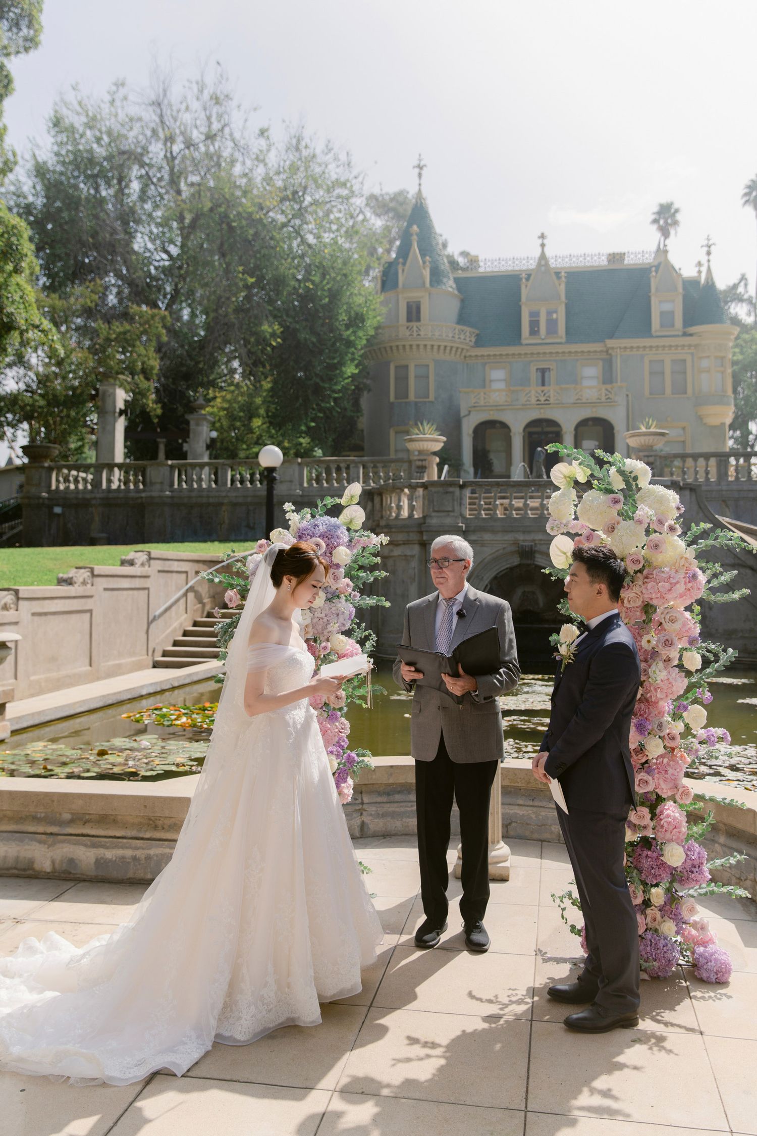 Outdoor wedding ceremony setup with pink floral arches at a castle-like venue.
