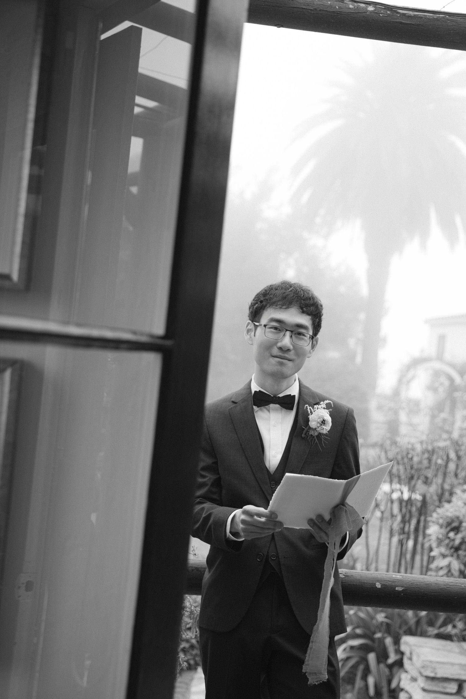 Person in black tuxedo reading from papers in an indoor venue.