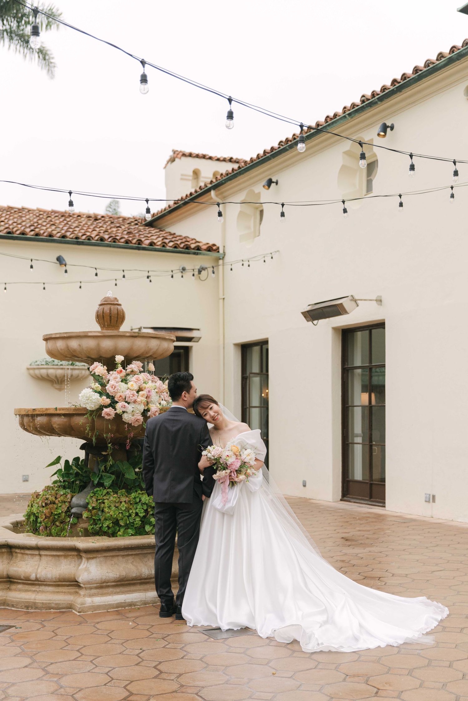 A couple poses in formal wedding attire in a Spanish courtyard with string lights overhead.