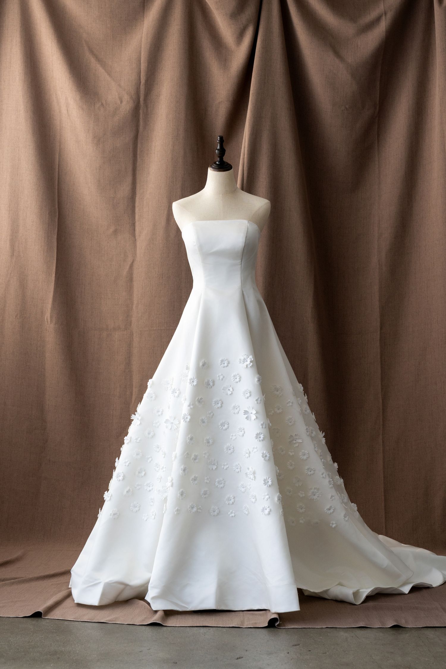 Full-length white wedding gown with floral details displayed on mannequin against brown background.