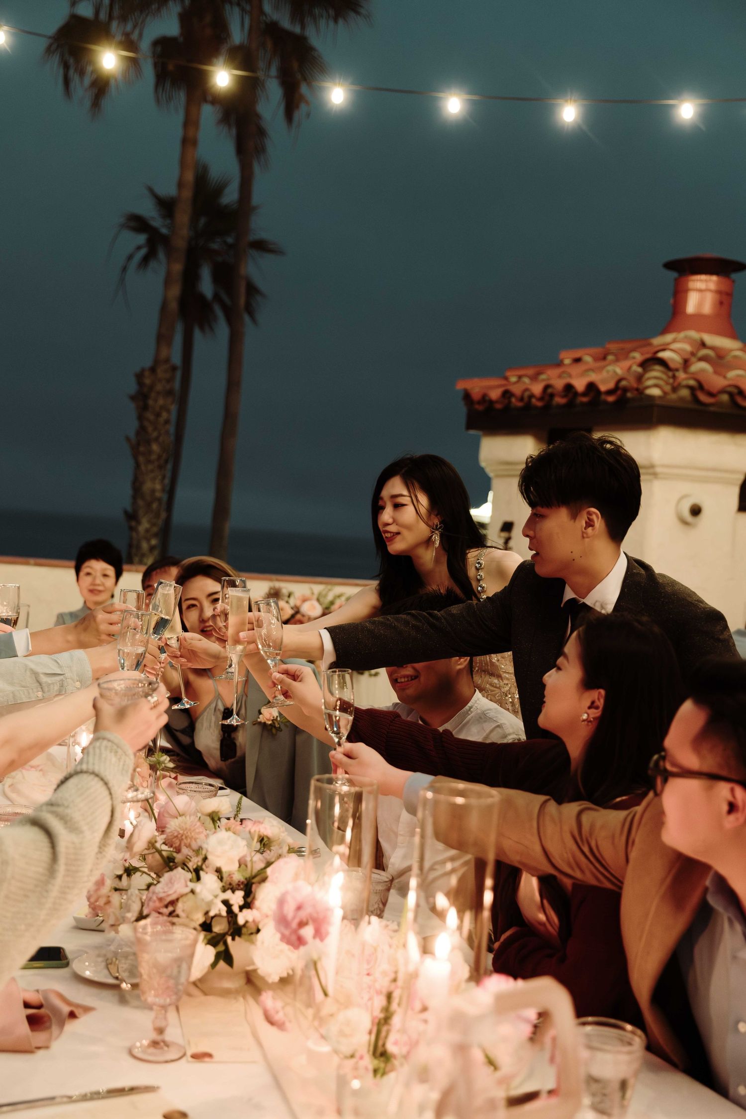 Wedding guests raising glasses for a toast under string lights at dusk.