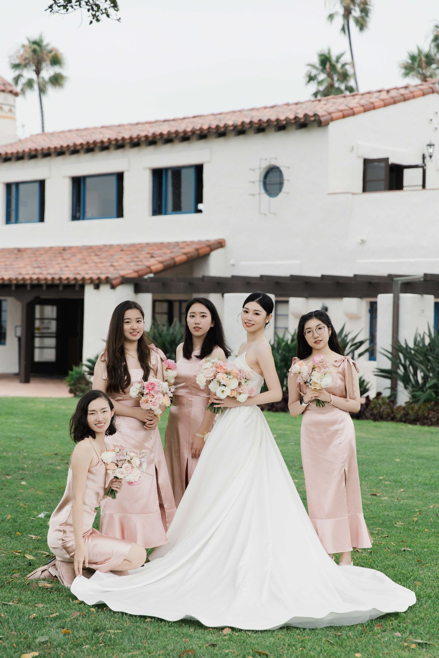 Wedding party in pink dresses gathered on grass near Spanish-style building.