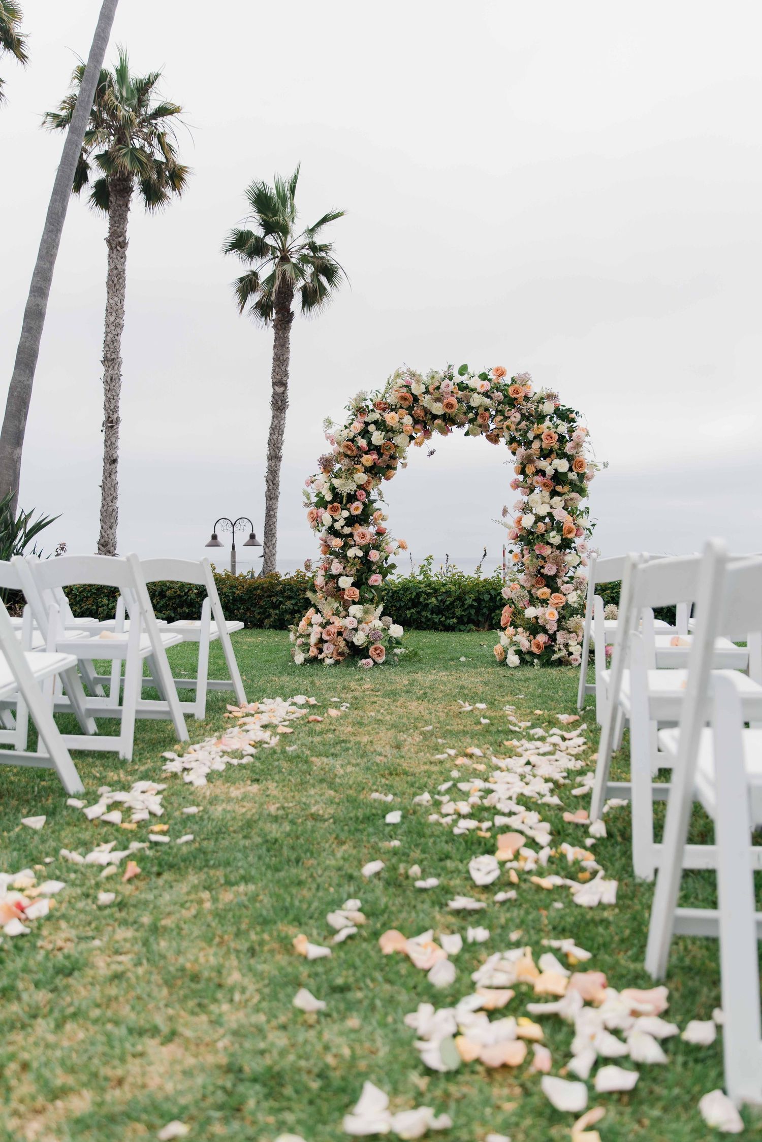Outdoor wedding ceremony setup with white chairs, floral arches, and palm trees along a grassy oceanfront venue.