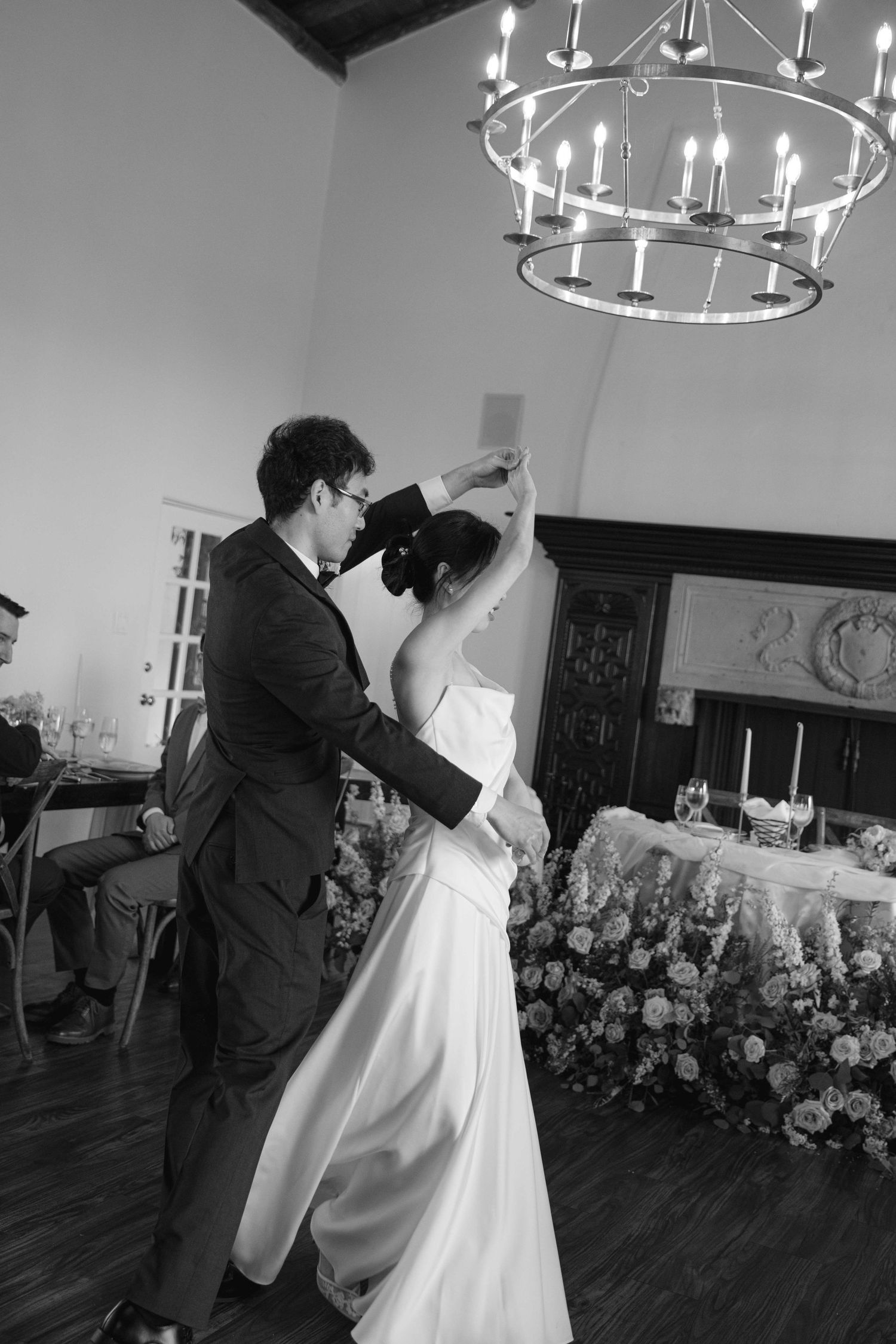 Black and white photo of newlyweds dancing under ornate chandeliers.
