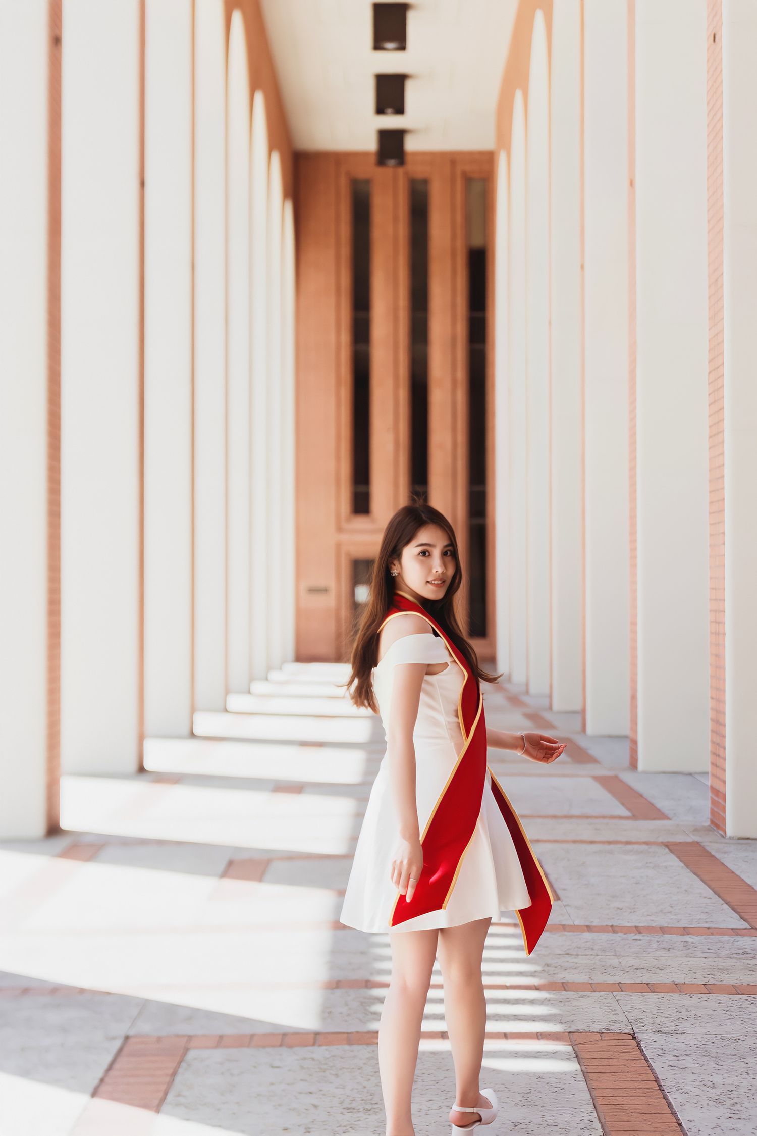 Graduate in red stole poses in a white columned hallway with dramatic lighting and architectural details.
