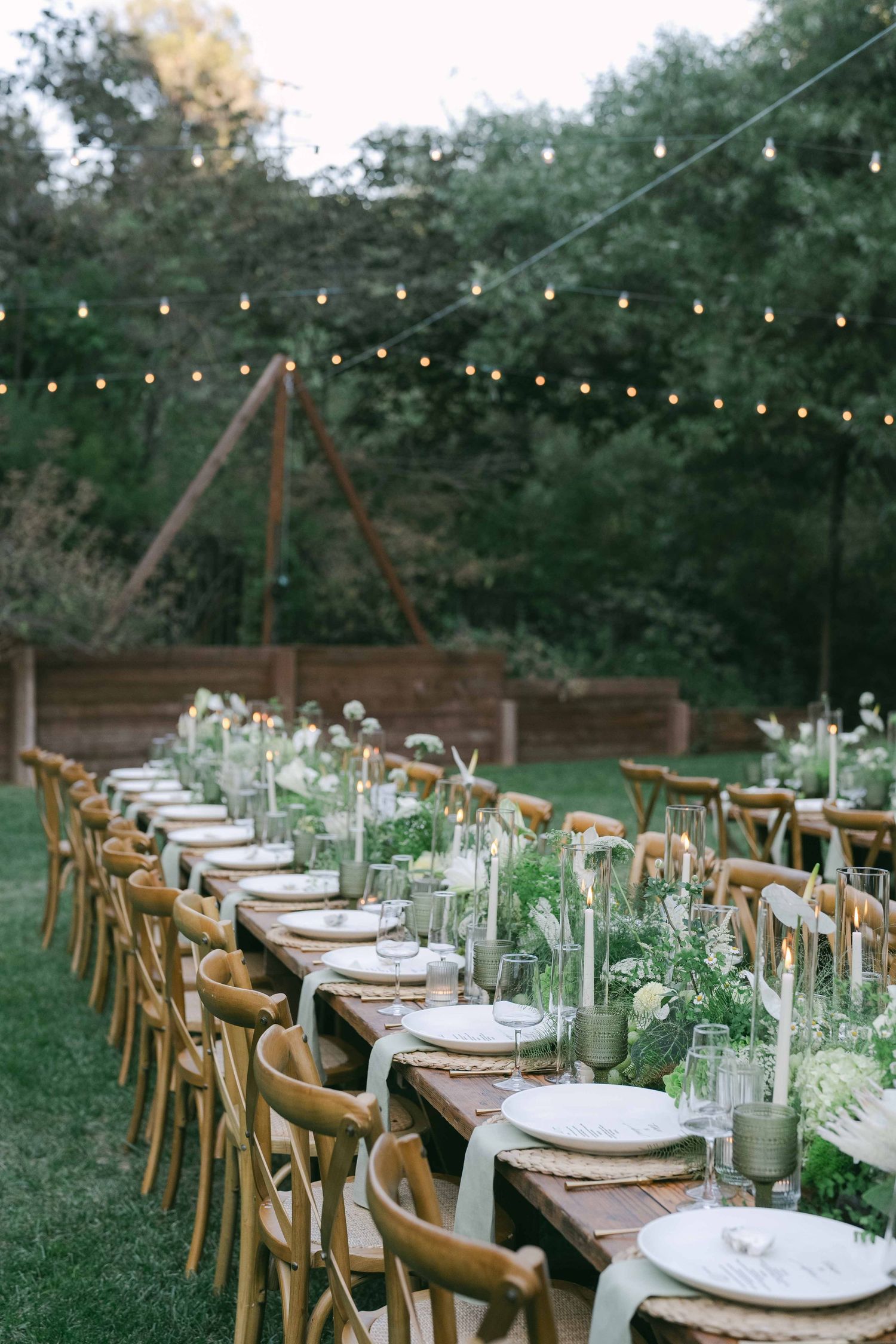 Long wooden dining table set for wedding reception with string lights and cross-back chairs.