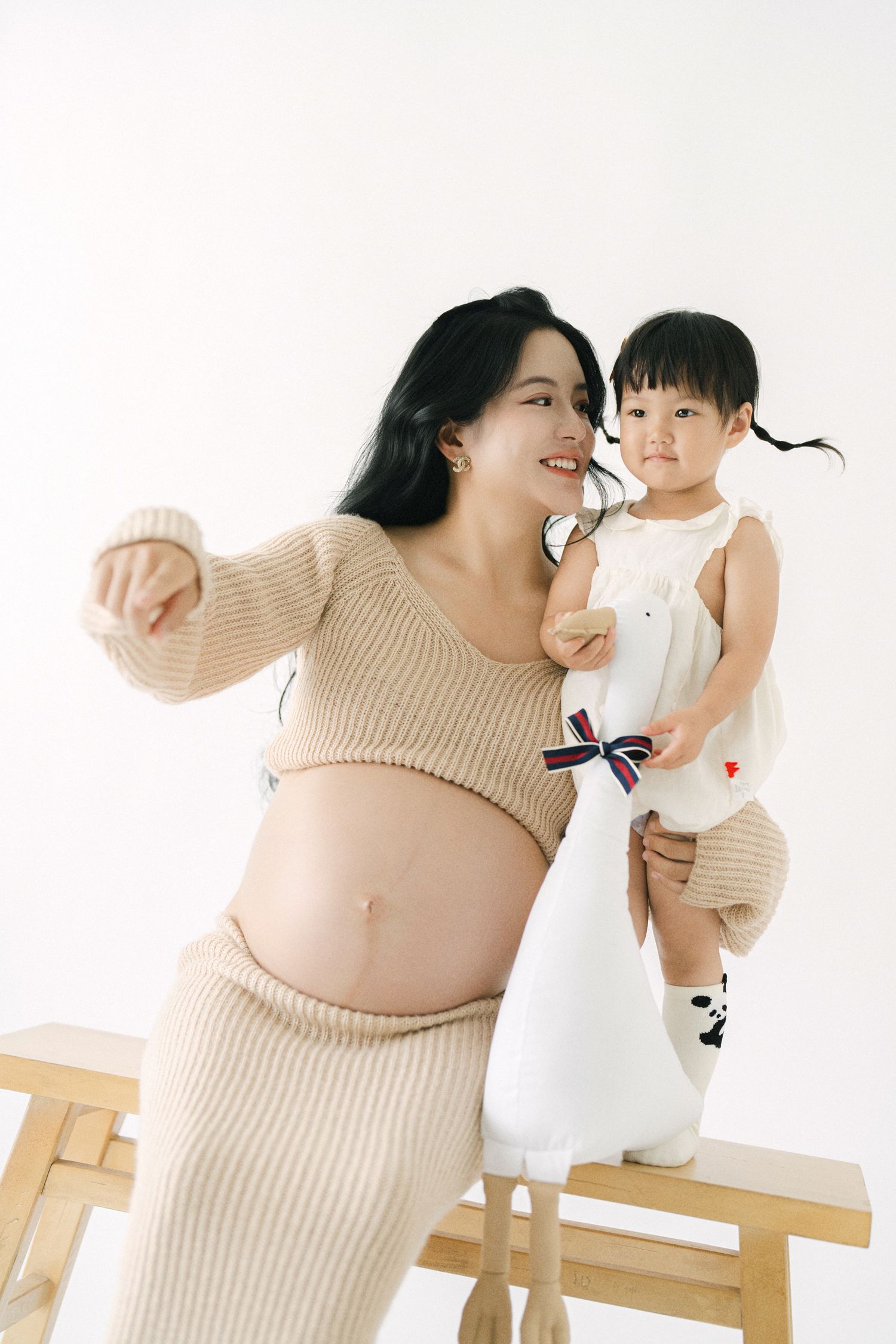 A pregnant woman in a beige sweater poses with a young child in a white dress against a white background.