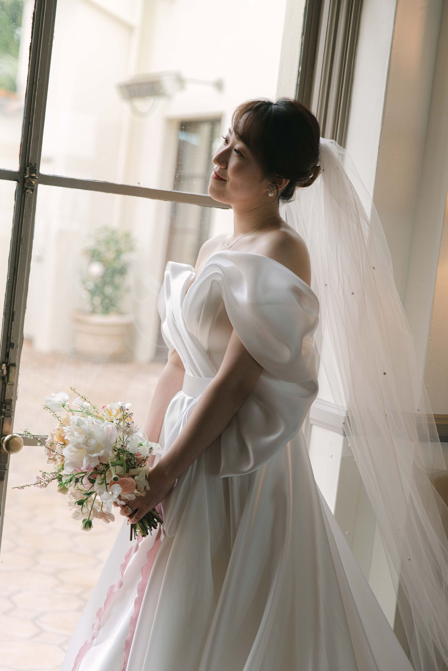 A person in a white wedding gown poses near large windows with a floral bouquet.
