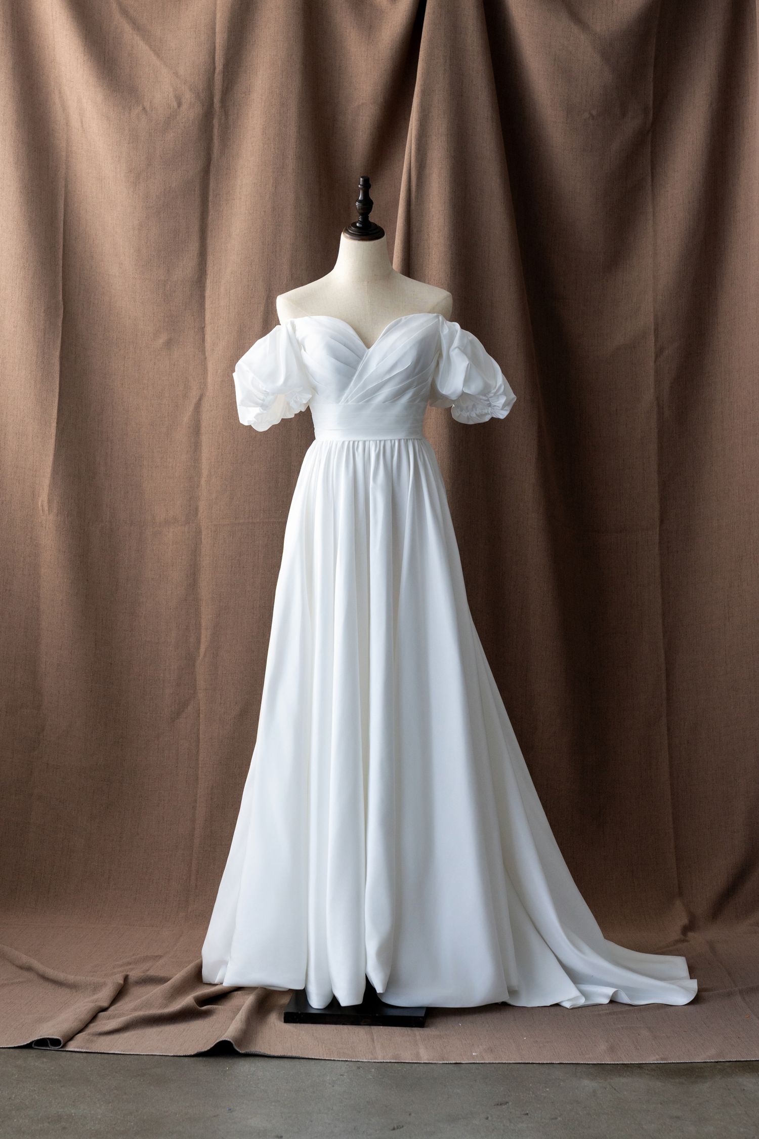 Flowing empire waist wedding gown with off-shoulder sleeves displayed against brown backdrop.