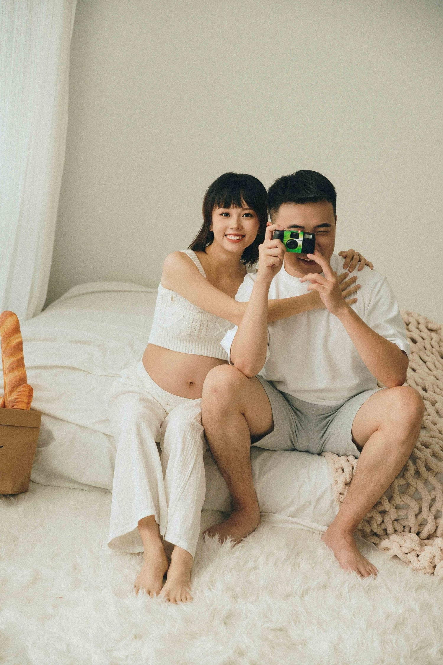 Couple lounging on white bed taking selfies and enjoying casual moments together.