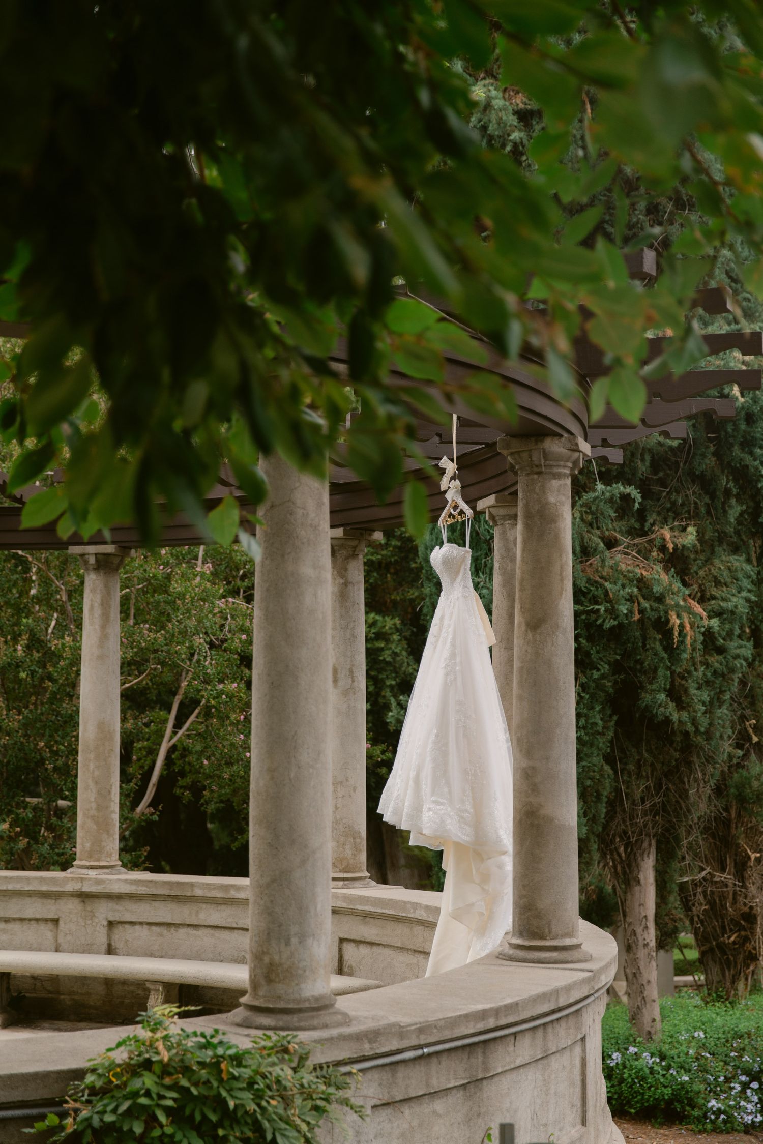 A white wedding dress hangs elegantly between classic stone columns in a garden setting.