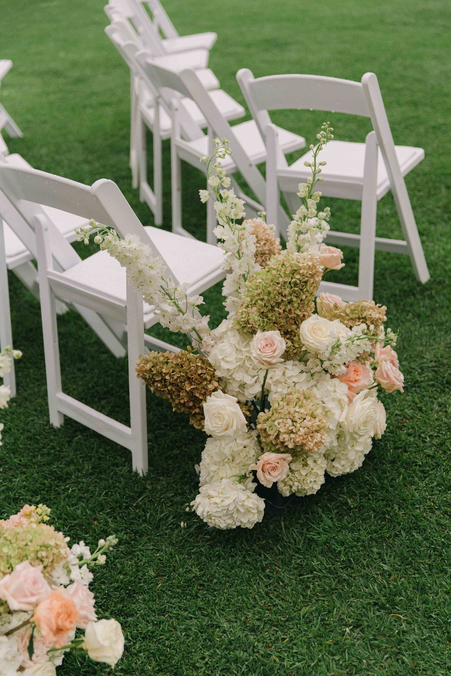 White floral arrangements with hydrangeas and roses decorate wedding ceremony aisle on green lawn.