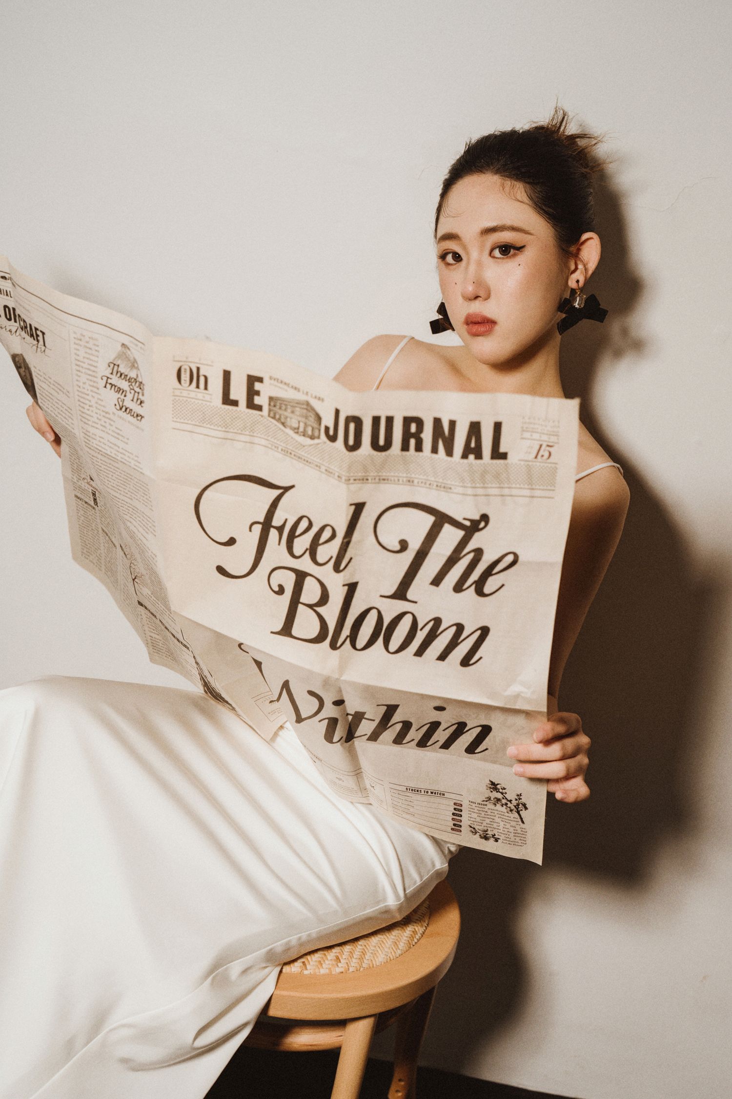 Fashion model in white dress poses with vintage newspaper against neutral backdrop in artistic photoshoot.
