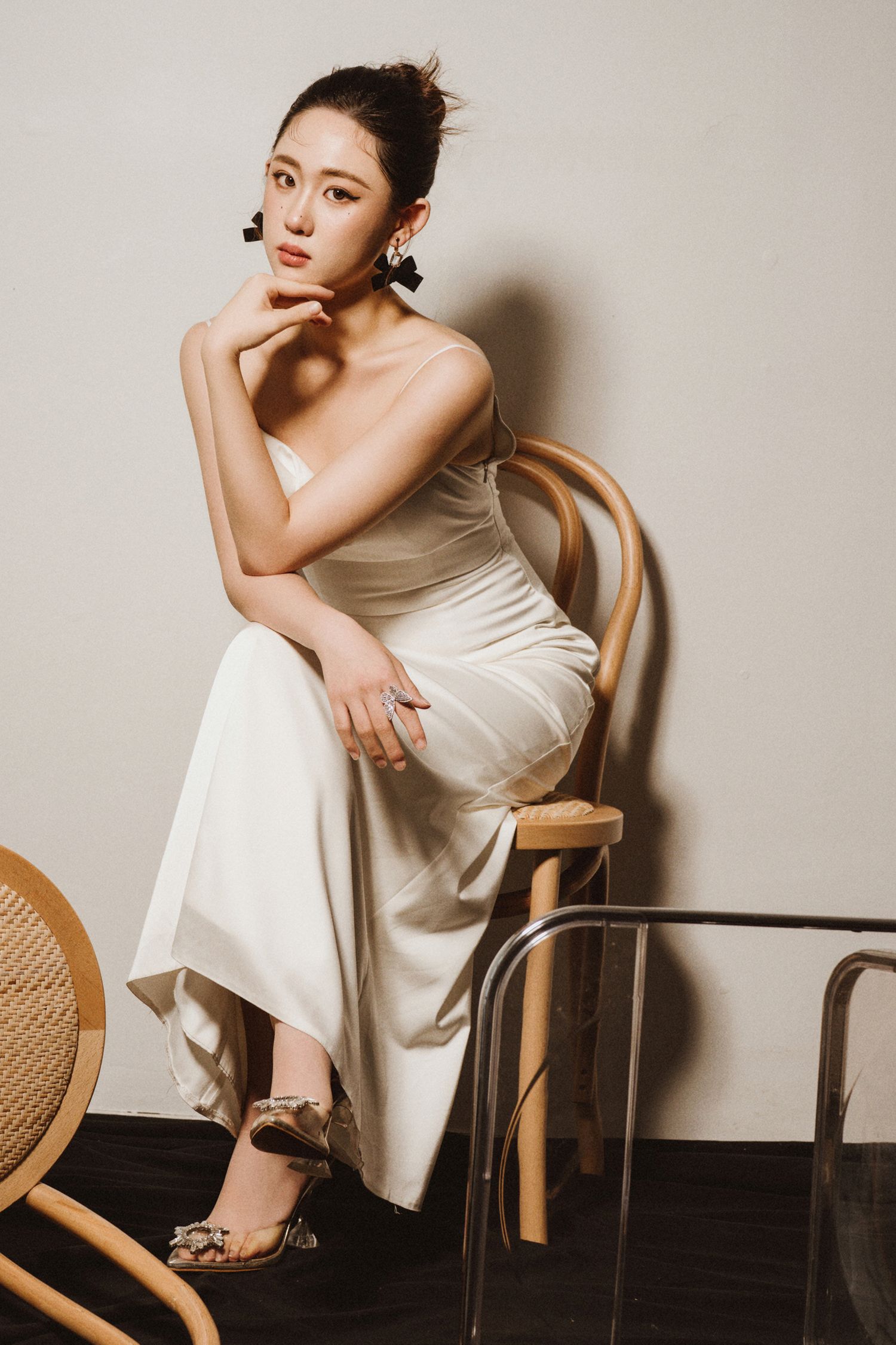 Model wearing sleeveless white dress sits elegantly on wooden chair in studio setting.