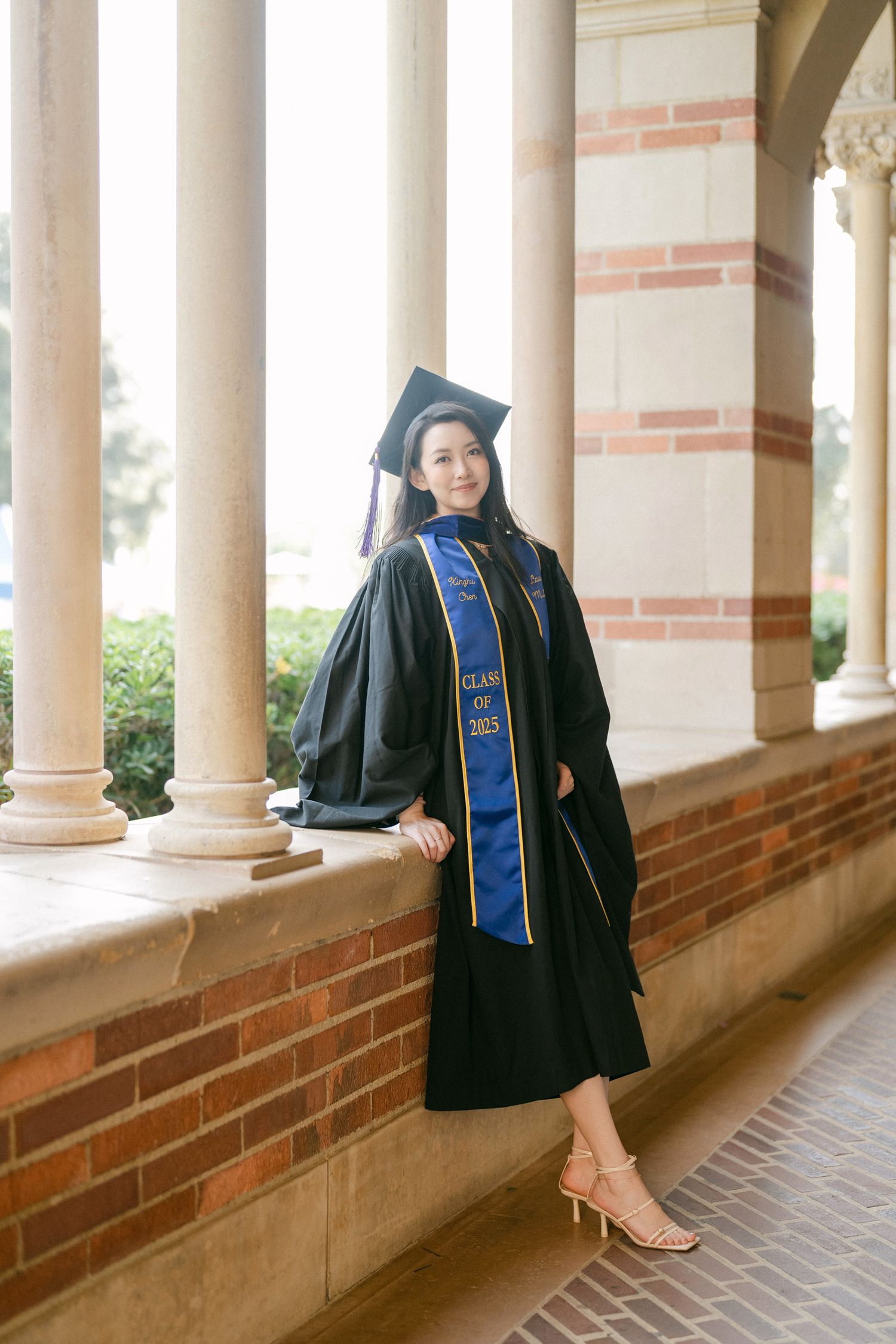 Graduate in black gown and blue stole poses along columned walkway of brick building.