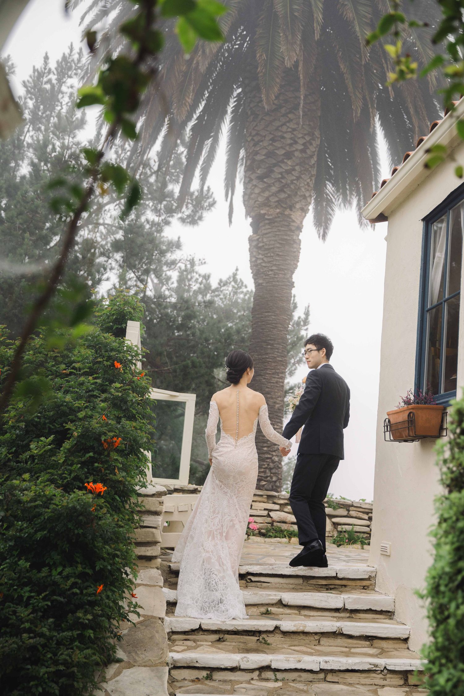 A couple in wedding attire stands on stone steps surrounded by lush greenery and palm trees on a misty day.