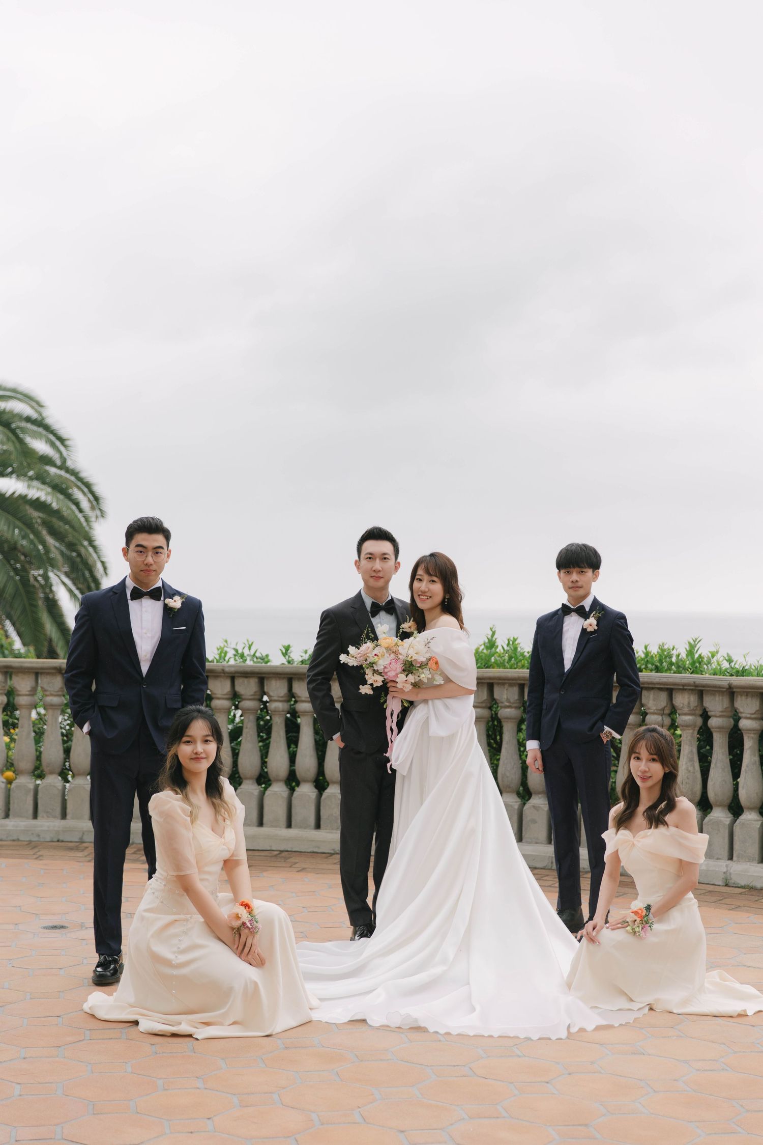 Wedding party poses together on an outdoor terrace with stone balustrade and palm trees.