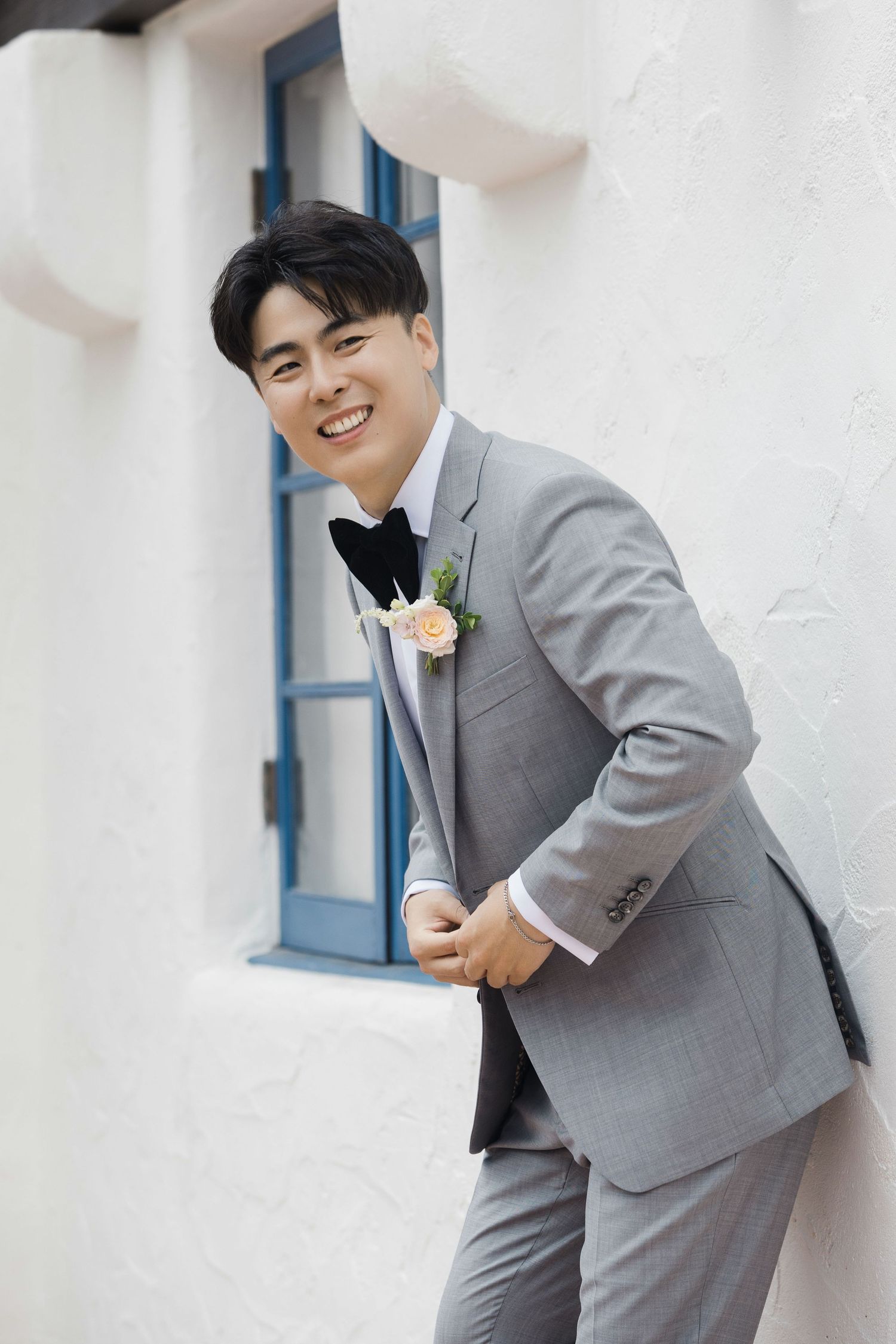 Groom in grey suit and black bowtie poses against white wall with blue door.