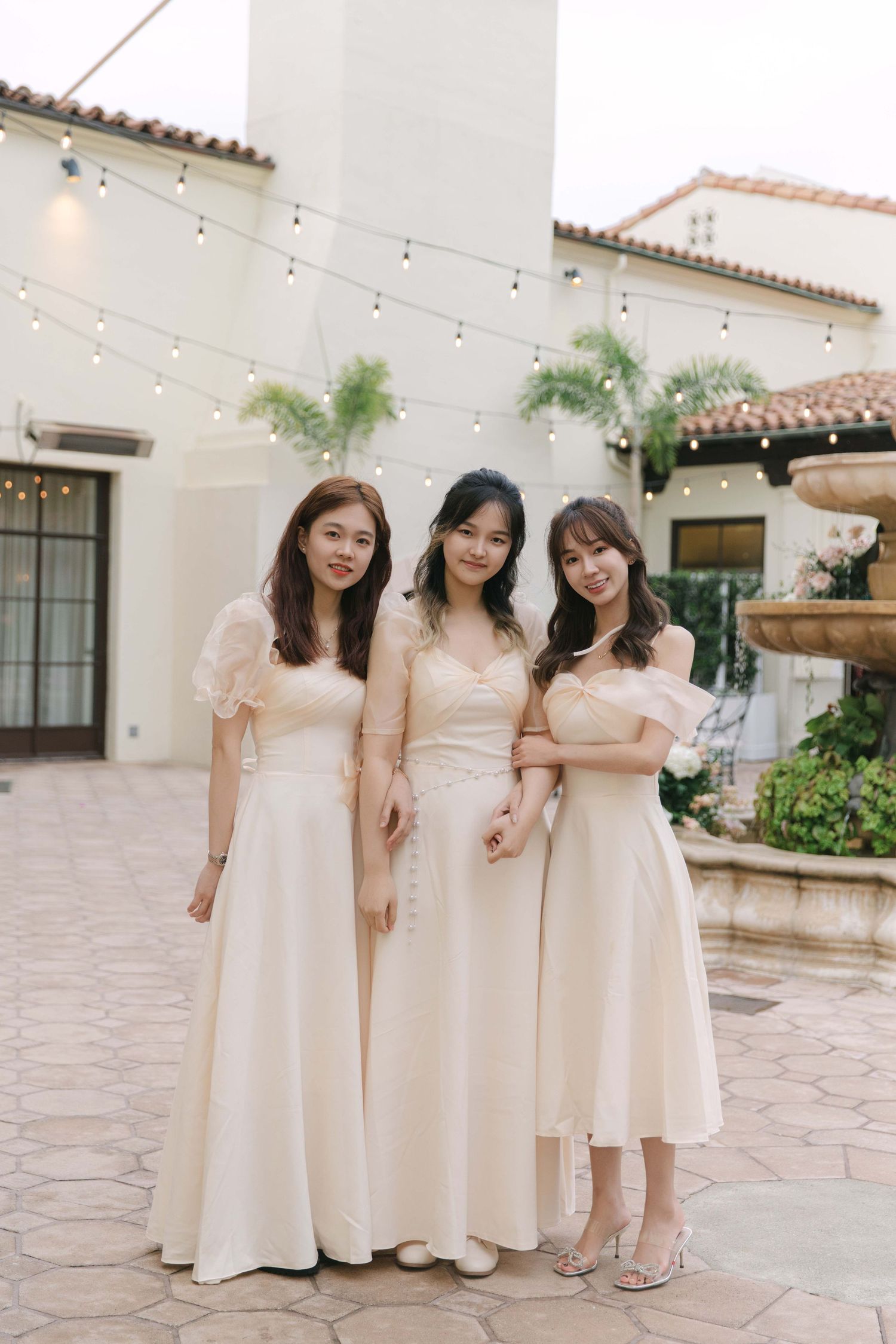 Three bridesmaids in blush pink dresses pose together on a stone patio with string lights.